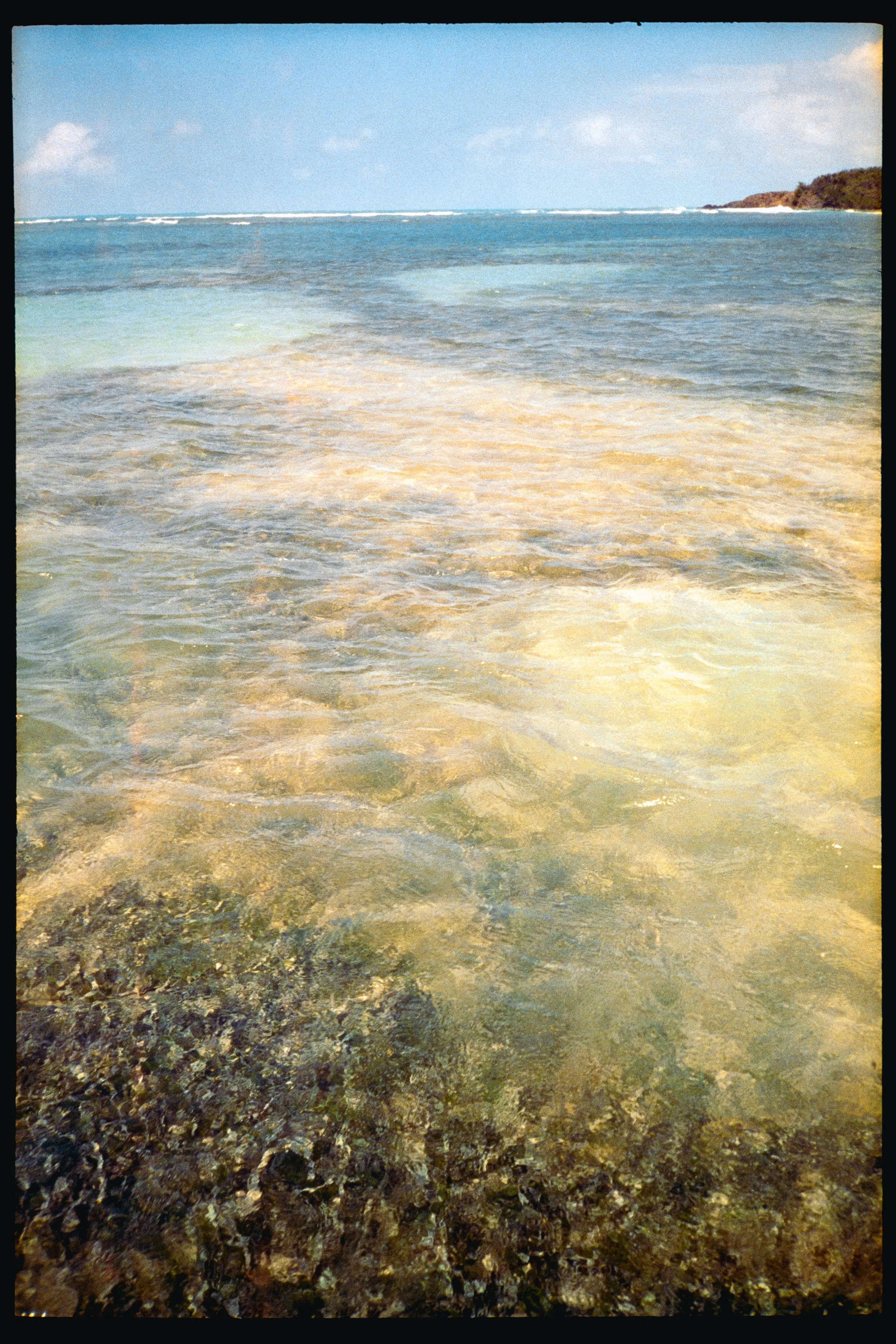 View of a shoreline with clear shallow water and small rocks at the beach, extending to a calm ocean under a partly cloudy sky, with a distant landmass on the horizon.