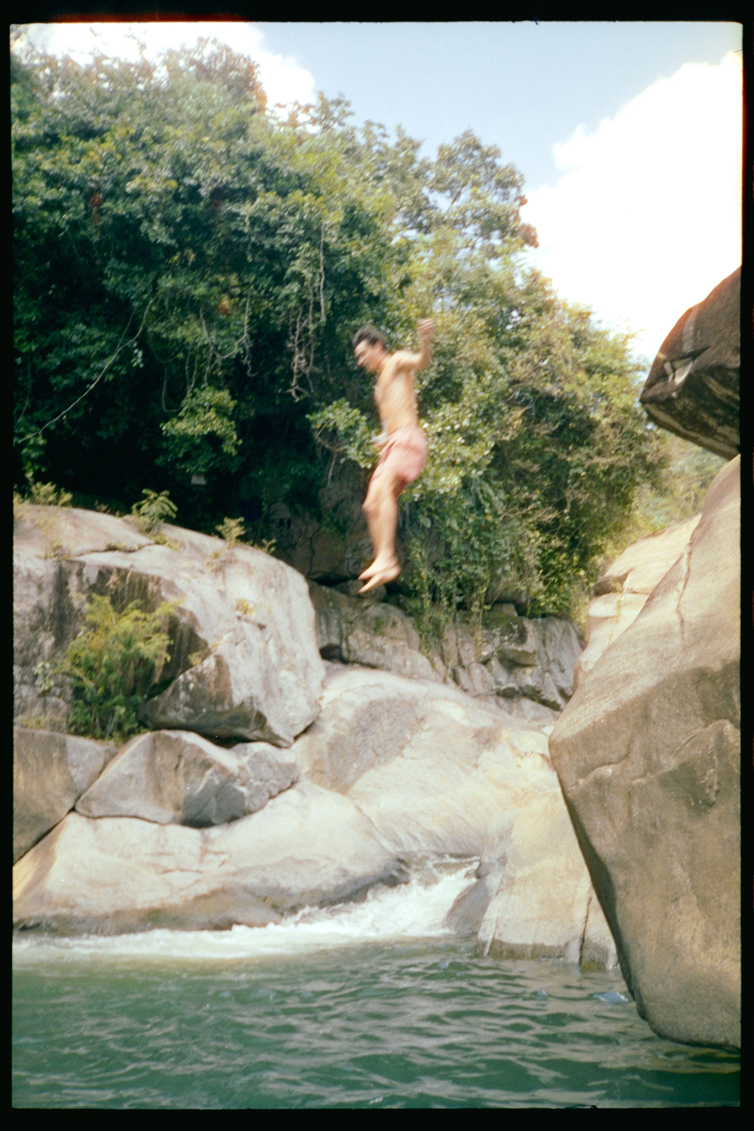 A person jumping from large rocks into a river with a rocky riverbank and trees in the background.