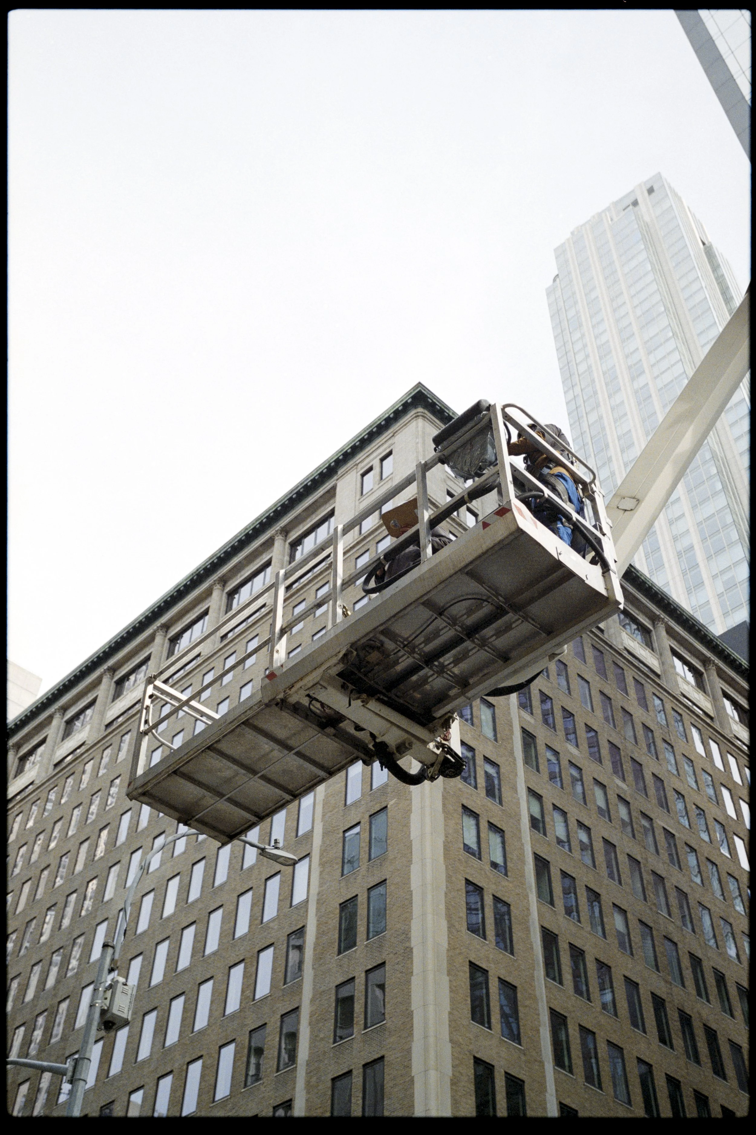 A worker in a safety harness on an elevated lift working on a building in an urban area with tall office buildings.