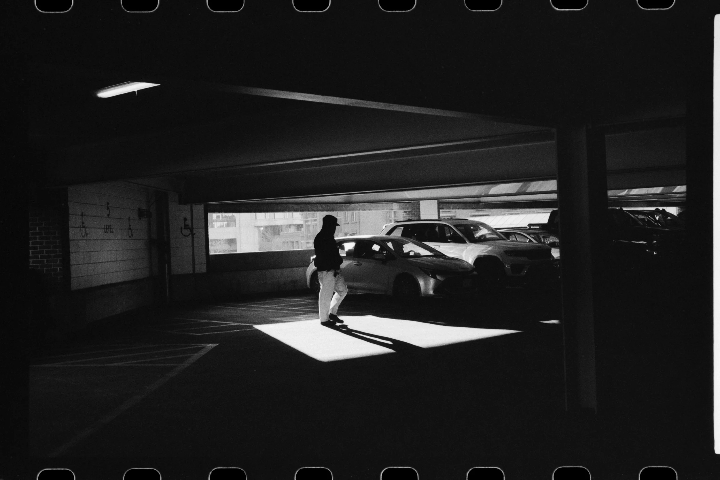 Black and white photo of a parking garage with cars parked and the silhouette of a man walking. Bright sunlight creates stark shadows on the ground.