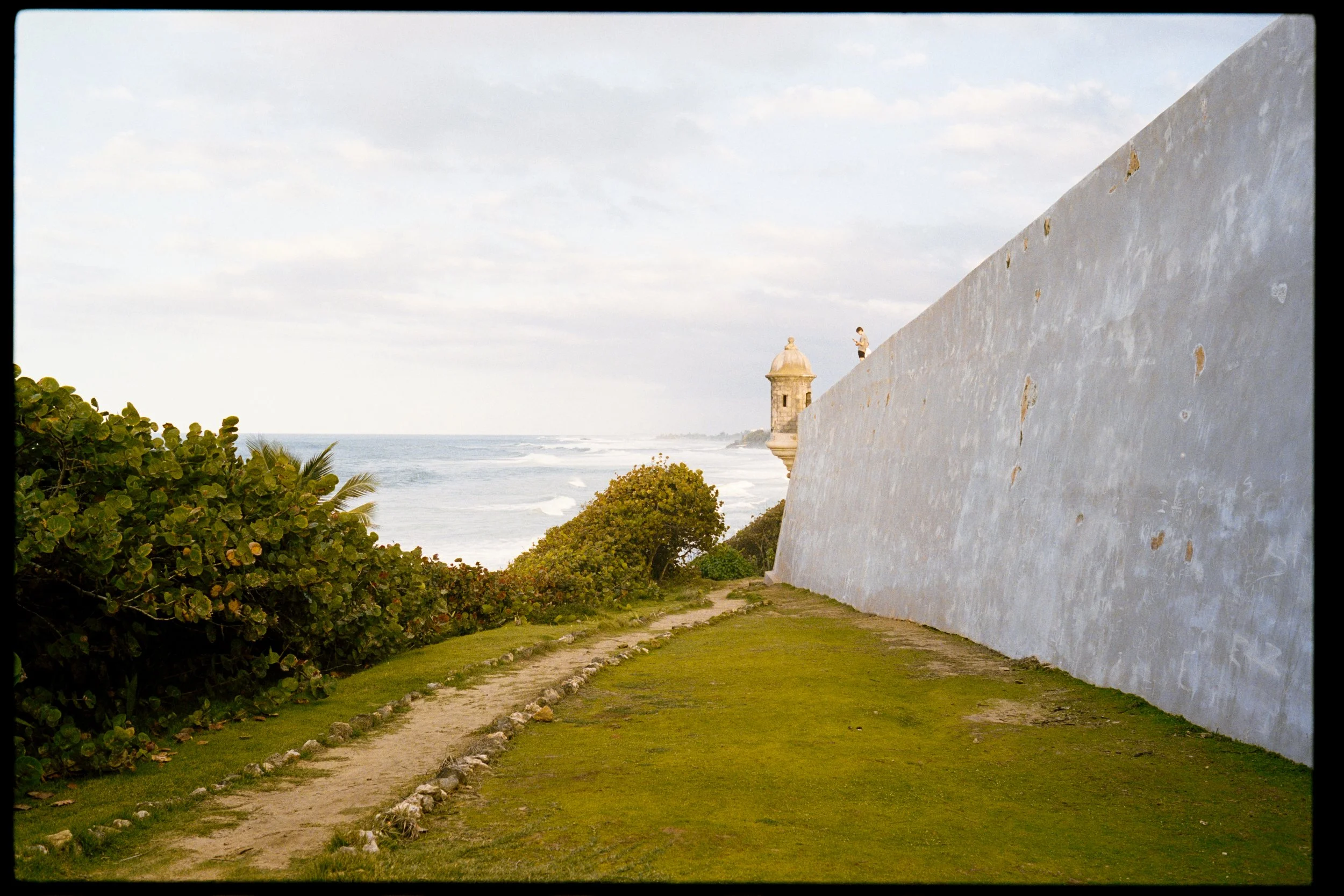 A coastal scene with a large white wall on the right, a pathway lined with rocks, lush green bushes on the left, and the ocean with waves and a cloudy sky in the background.