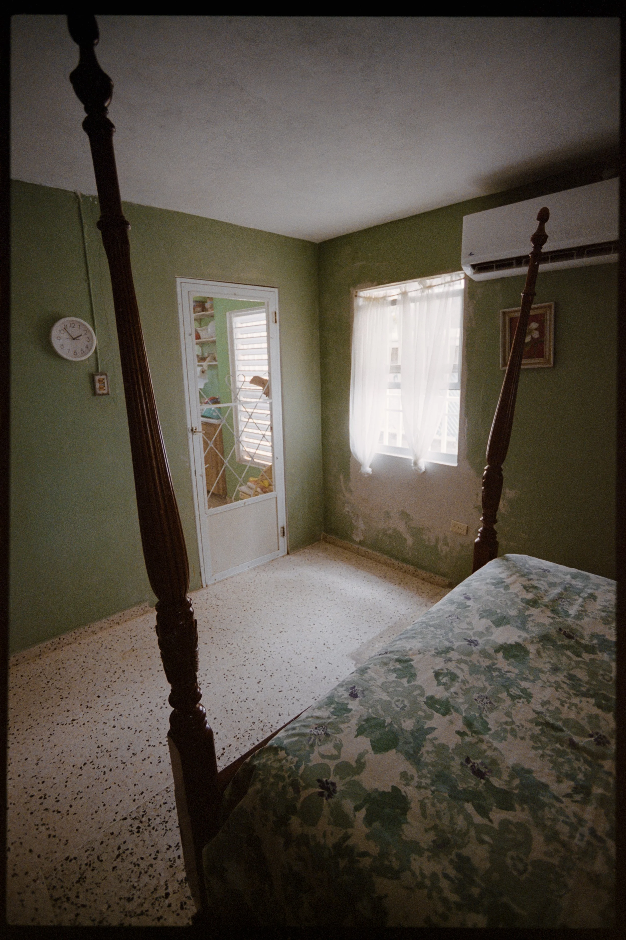A bedroom with green walls, a window with white curtains, an air conditioning unit, a floral bedspread, a clock on the wall, and a door with a glass panel leading outside. The bed has a dark wooden four-poster frame.