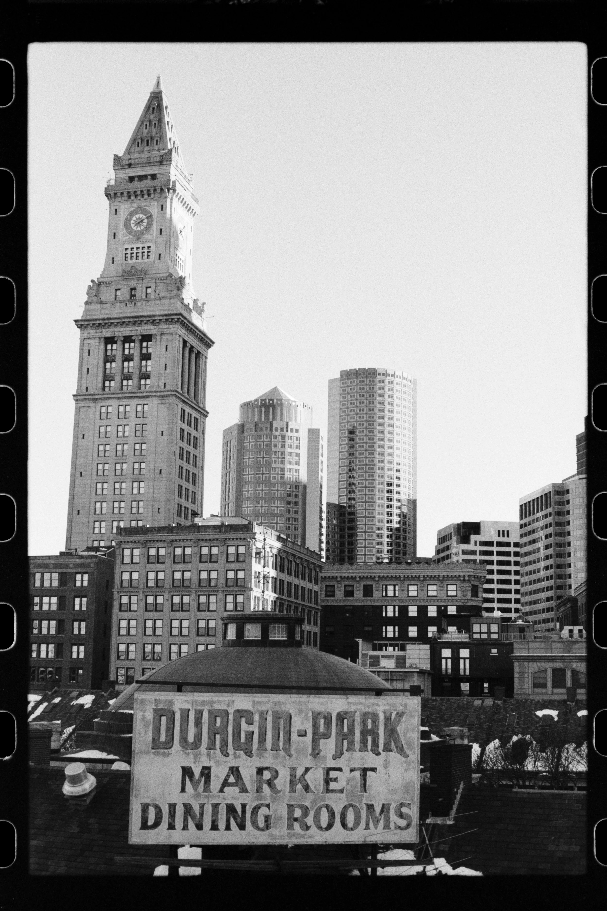 Black and white photograph of a city skyline with tall buildings, including the iconic clock tower, and a sign in the foreground reading "Durham-Park Market, Diner, Deli, Docks."