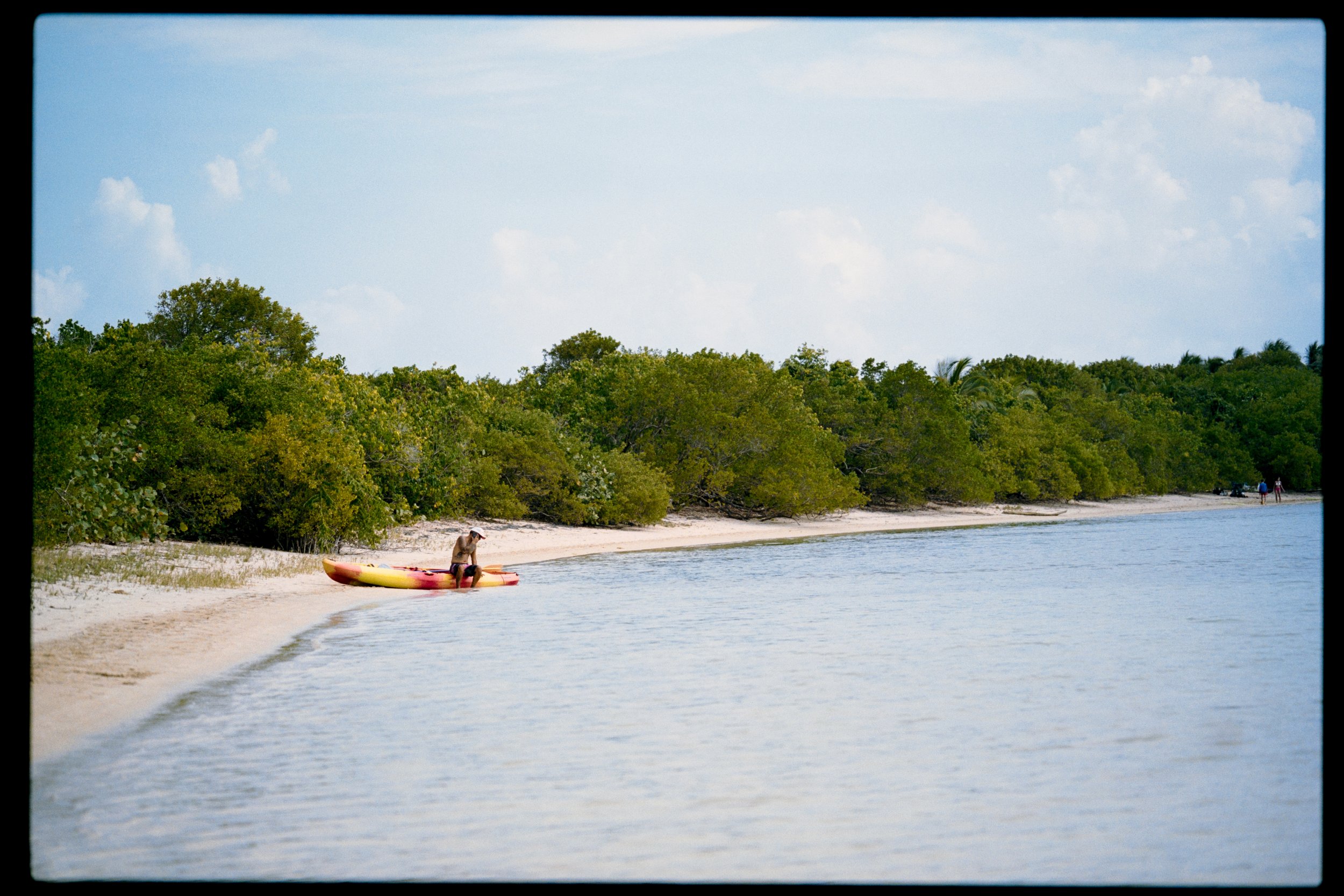 Person sitting on colorful kayak near the shoreline of a tropical beach with dense green foliage and a blue sky.