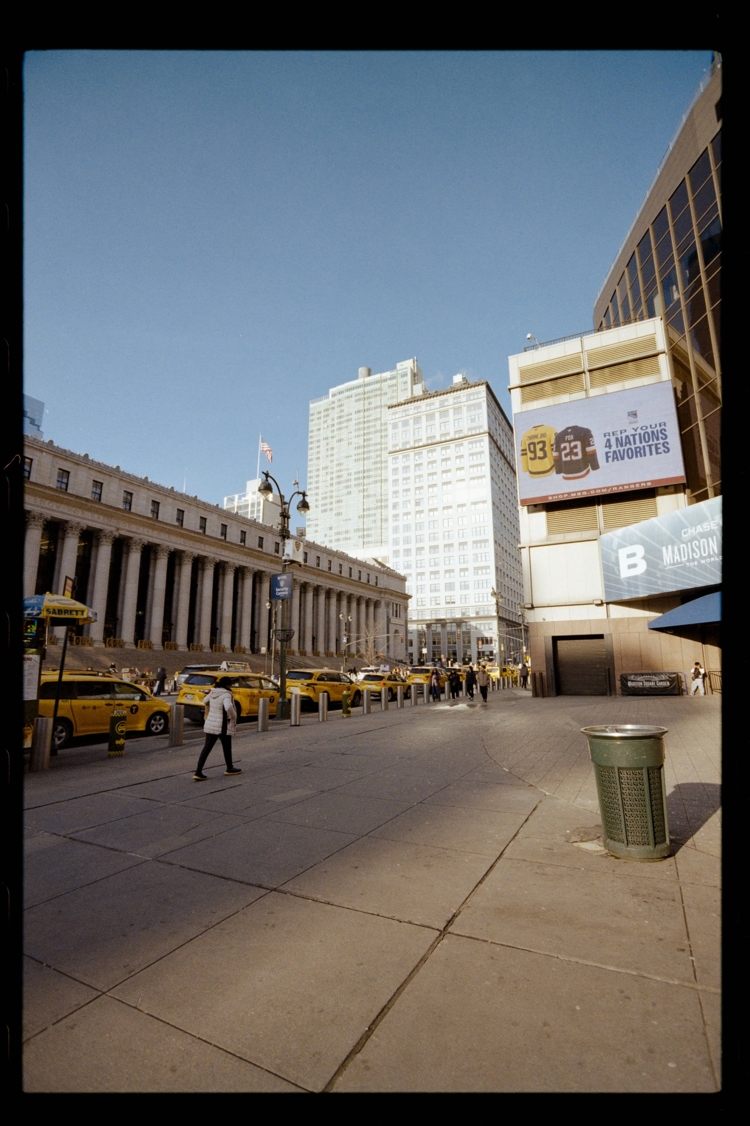 Urban street scene with yellow taxis, pedestrians walking, modern and historic buildings, and billboard advertisements, including one for sports jerseys, against a clear blue sky.