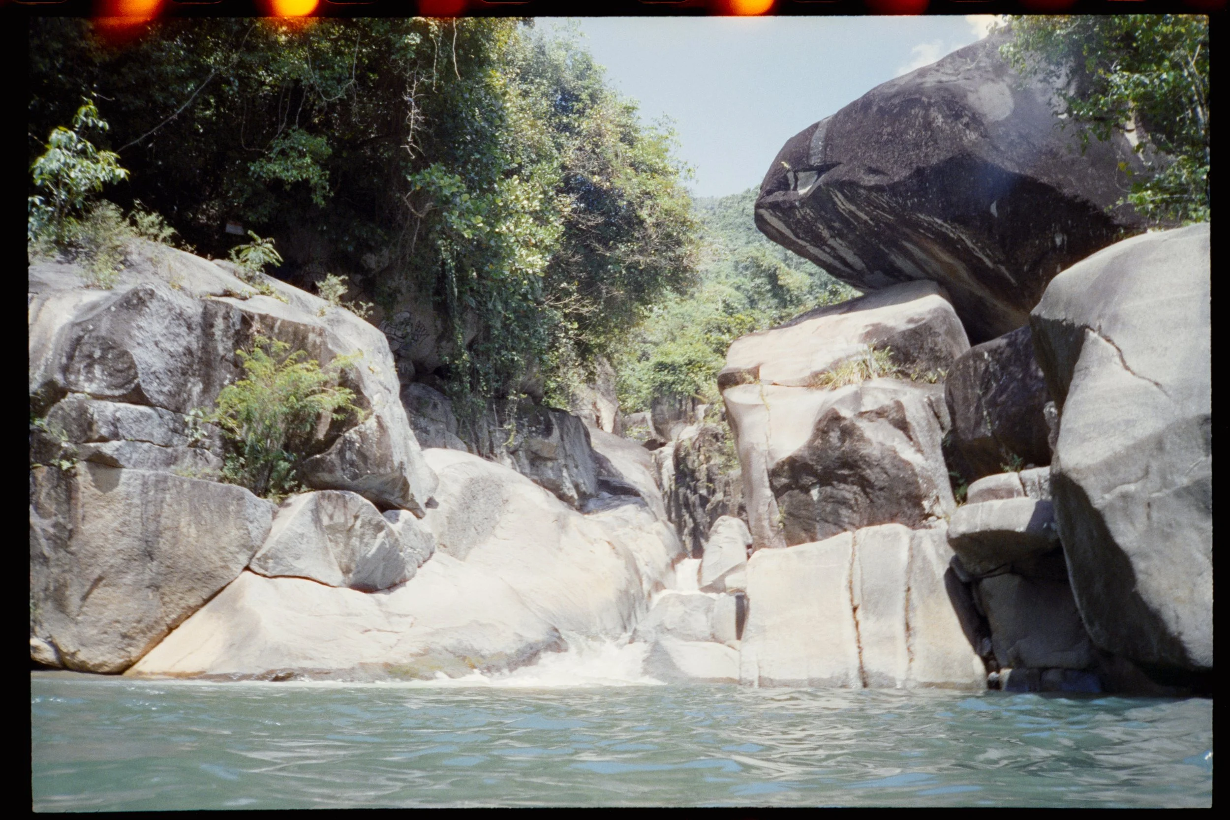 A rocky riverbed with large boulders and green foliage, with a river flowing at the bottom of the image and trees in the background.