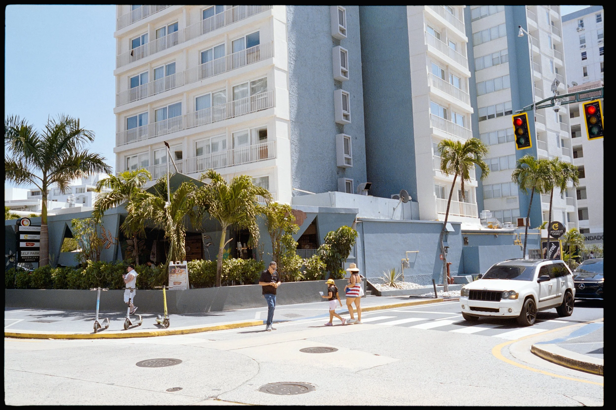 People walking on a sidewalk in front of a tall city building with multiple balconies and a cafe at street level, with cars parked and scooter rentals along the curb.
