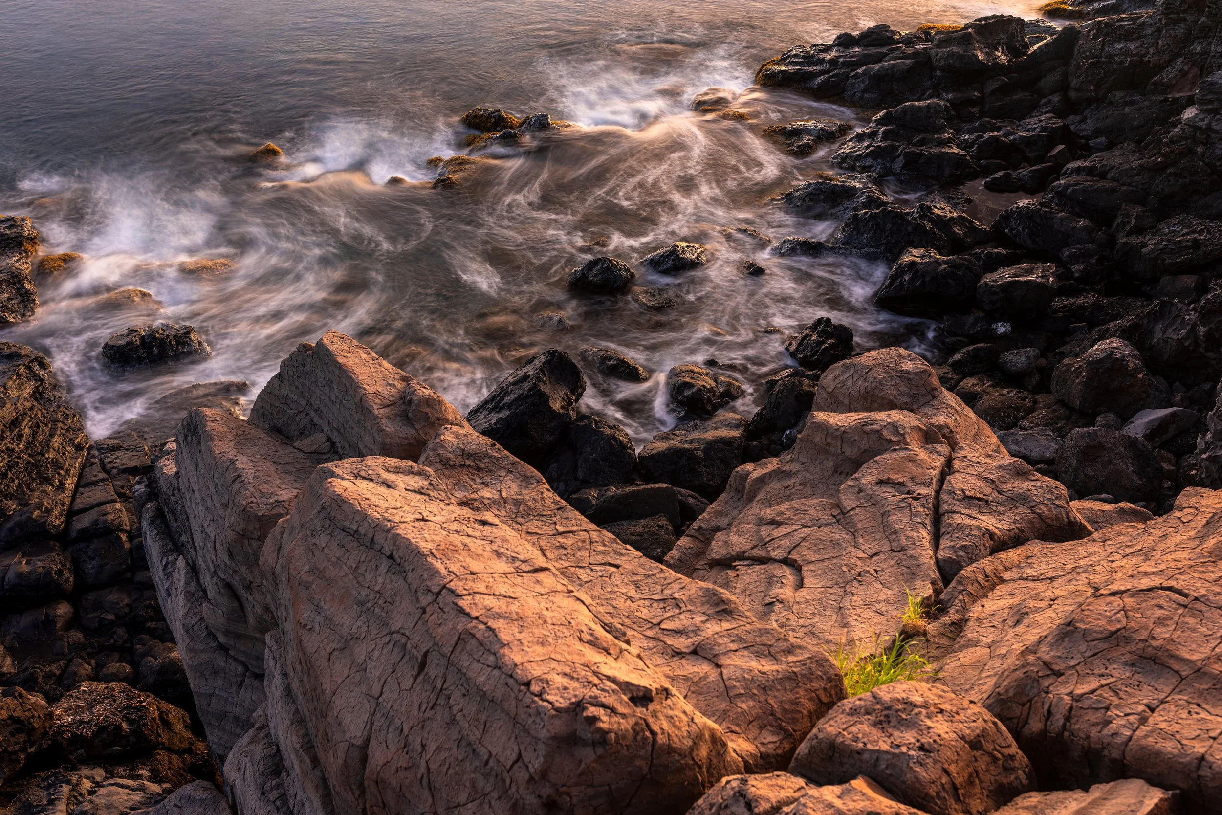 Rocks along a shoreline with waves gently washing over them at sunset.