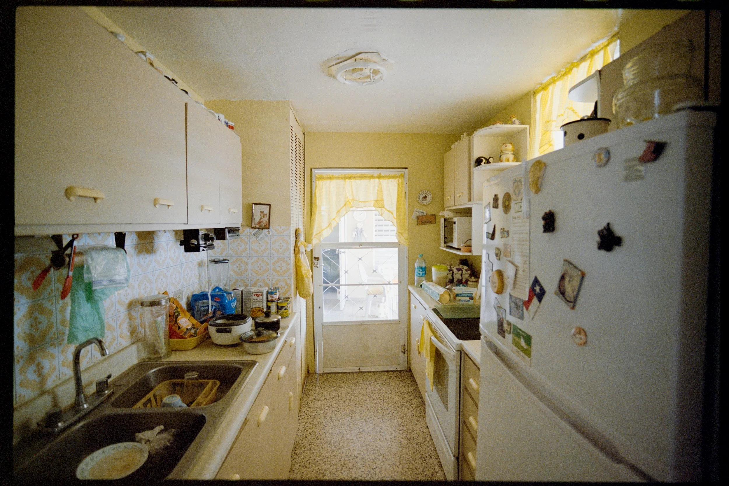 A small kitchen with yellow fixtures and cabinets, cluttered countertops, a refrigerator covered with magnets, a window with yellow curtains, and various kitchen appliances and items.