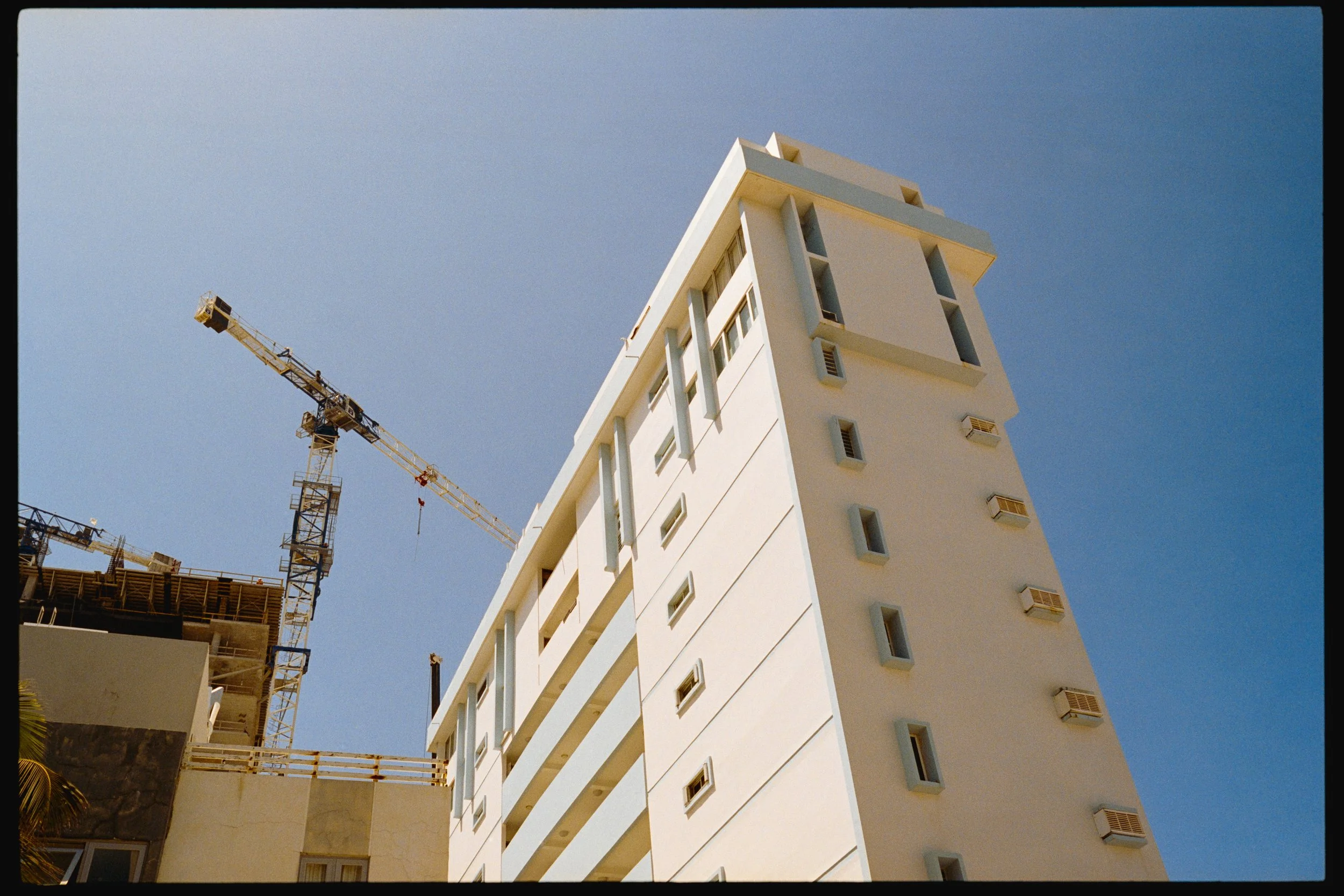 A tall, modern white building with multiple small rectangular windows, some with air conditioning units, under a clear blue sky. Construction cranes are visible to the left of the building.