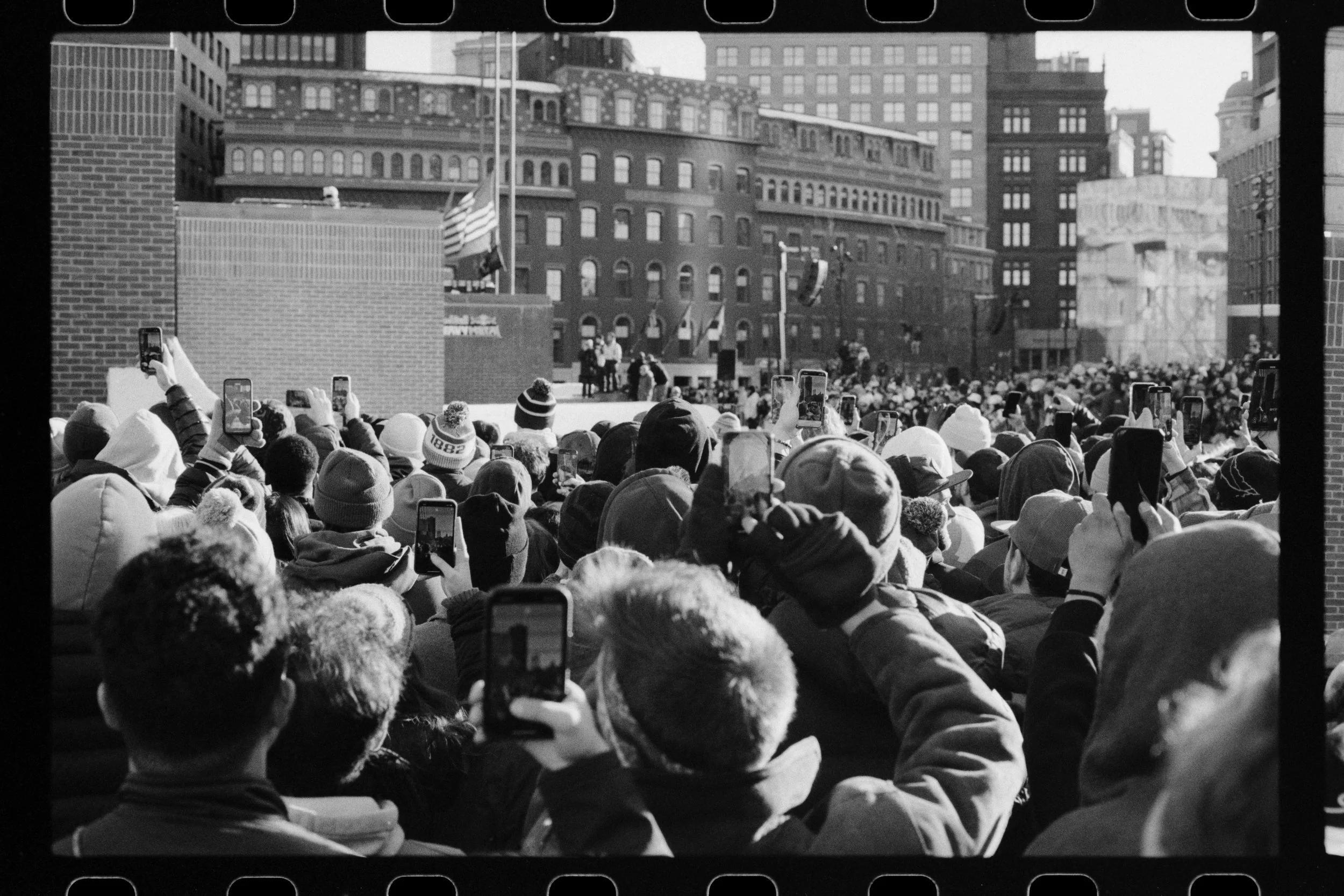 A large crowd of people wearing winter hats, many holding smartphones to take pictures, gather outdoors in an urban setting. There is a stage or platform with people talking or performing, with buildings in the background and American flags flying.