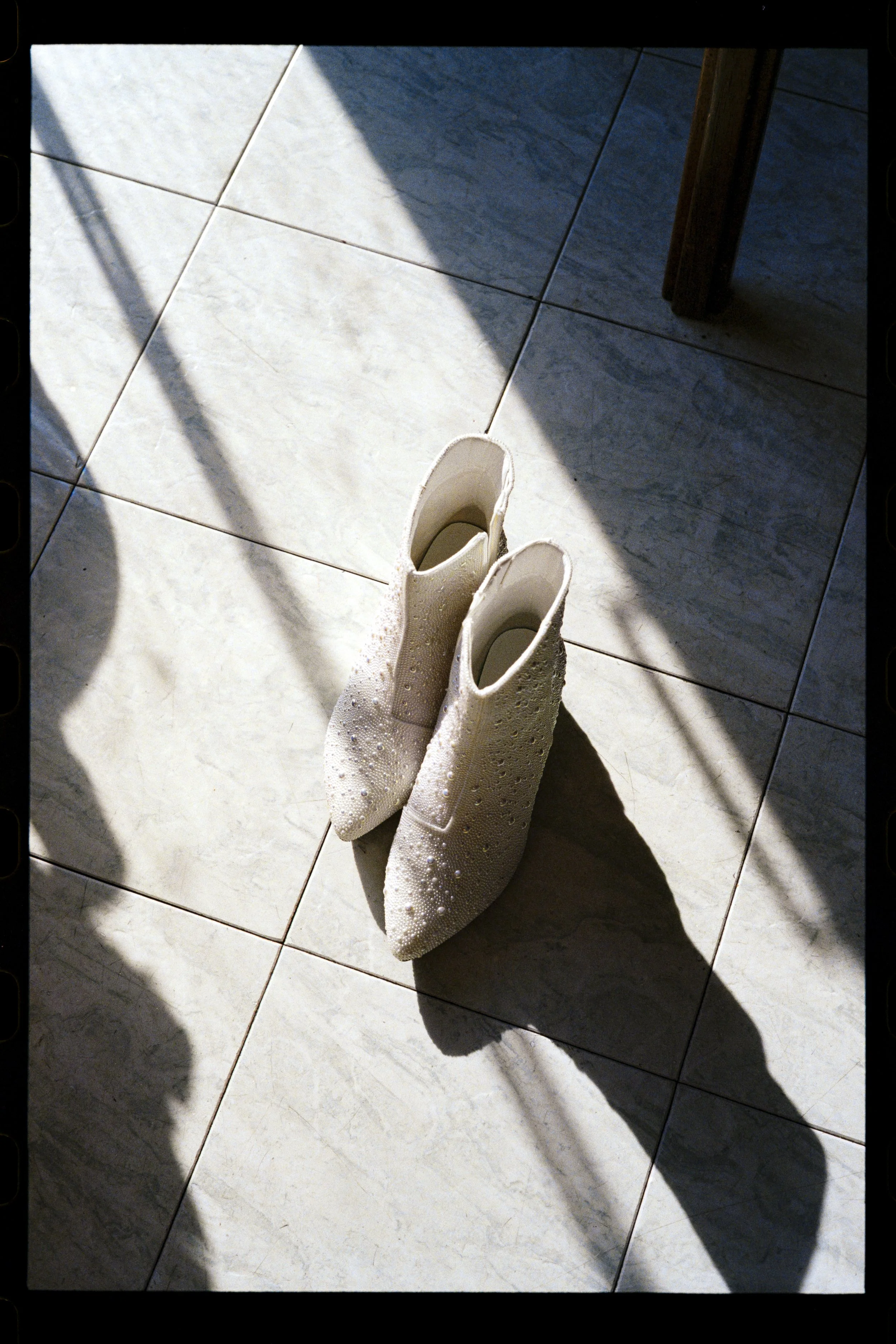 Pair of white high-heeled shoes with decorative embellishments on a tiled floor, with your shadow and a chair leg visible in the scene.