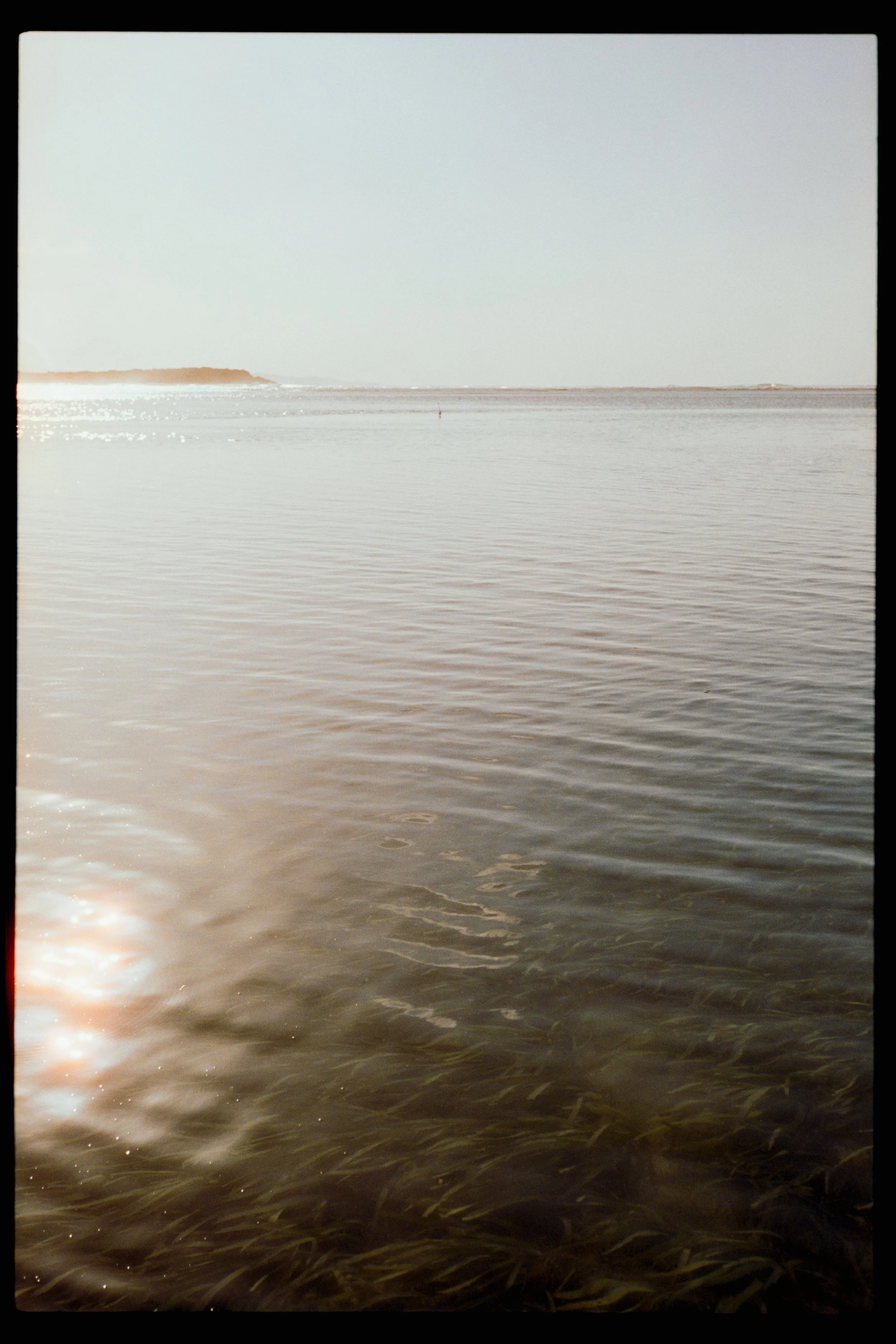 Sunset or sunrise over a calm body of water, with a distant shoreline visible on the horizon.
