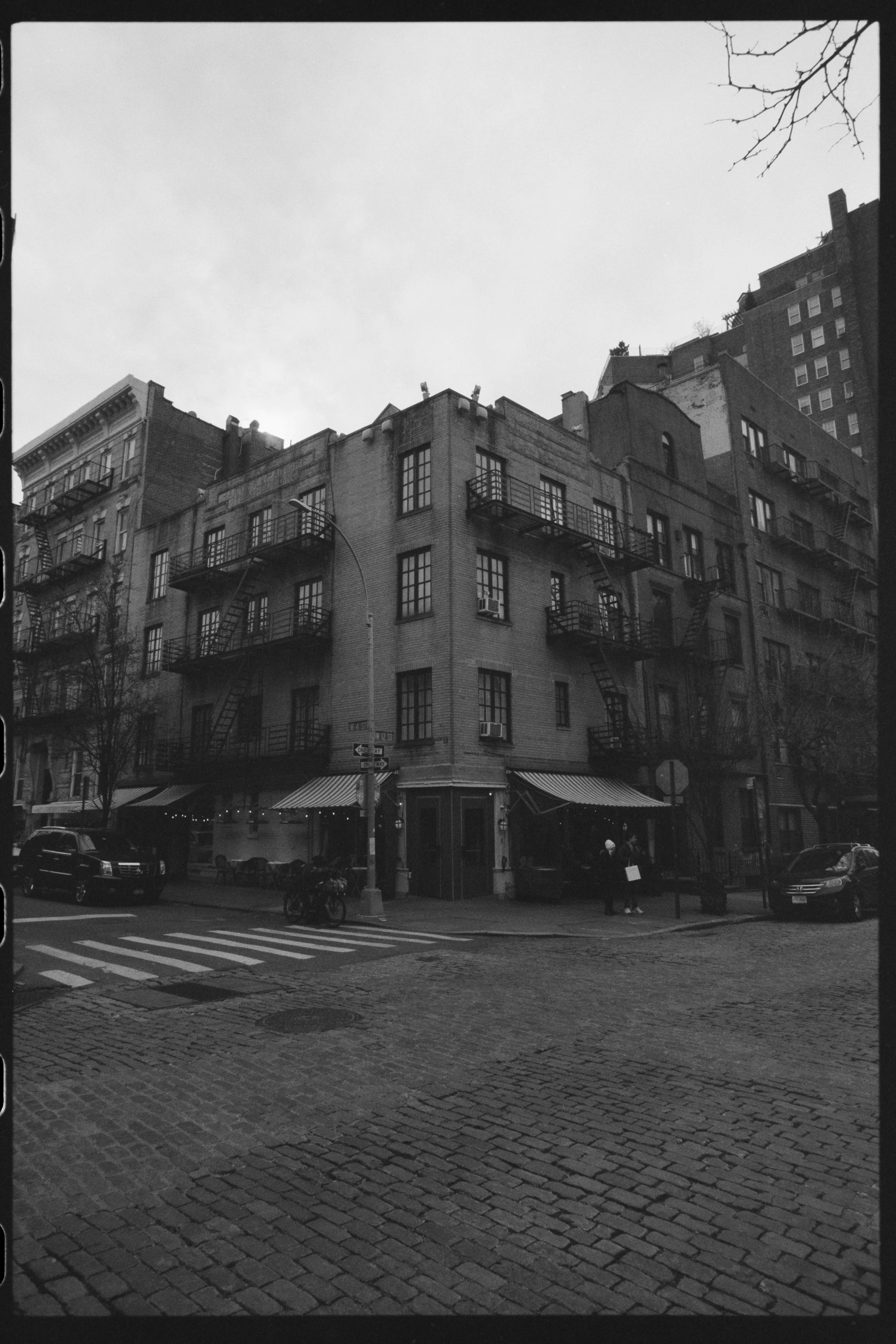 Nighttime street scene featuring a multi-story brick building with fire escapes, a storefront on the ground floor, parked cars, and a cobblestone crosswalk. Bare tree branches are visible in the top right corner.
