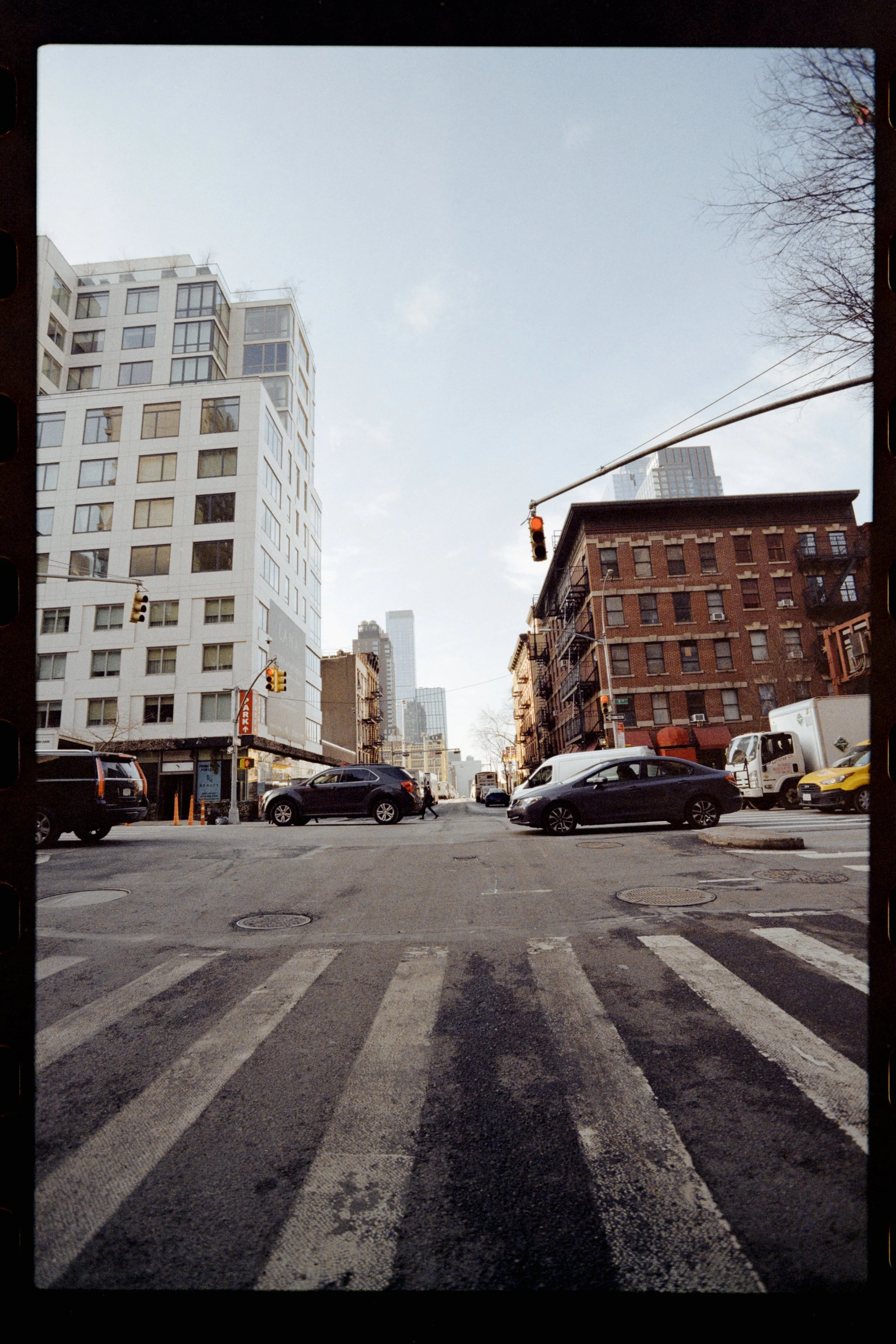 City street intersection with crosswalk, cars stopped at traffic lights, multi-story buildings, and leafless trees under a cloudy sky.