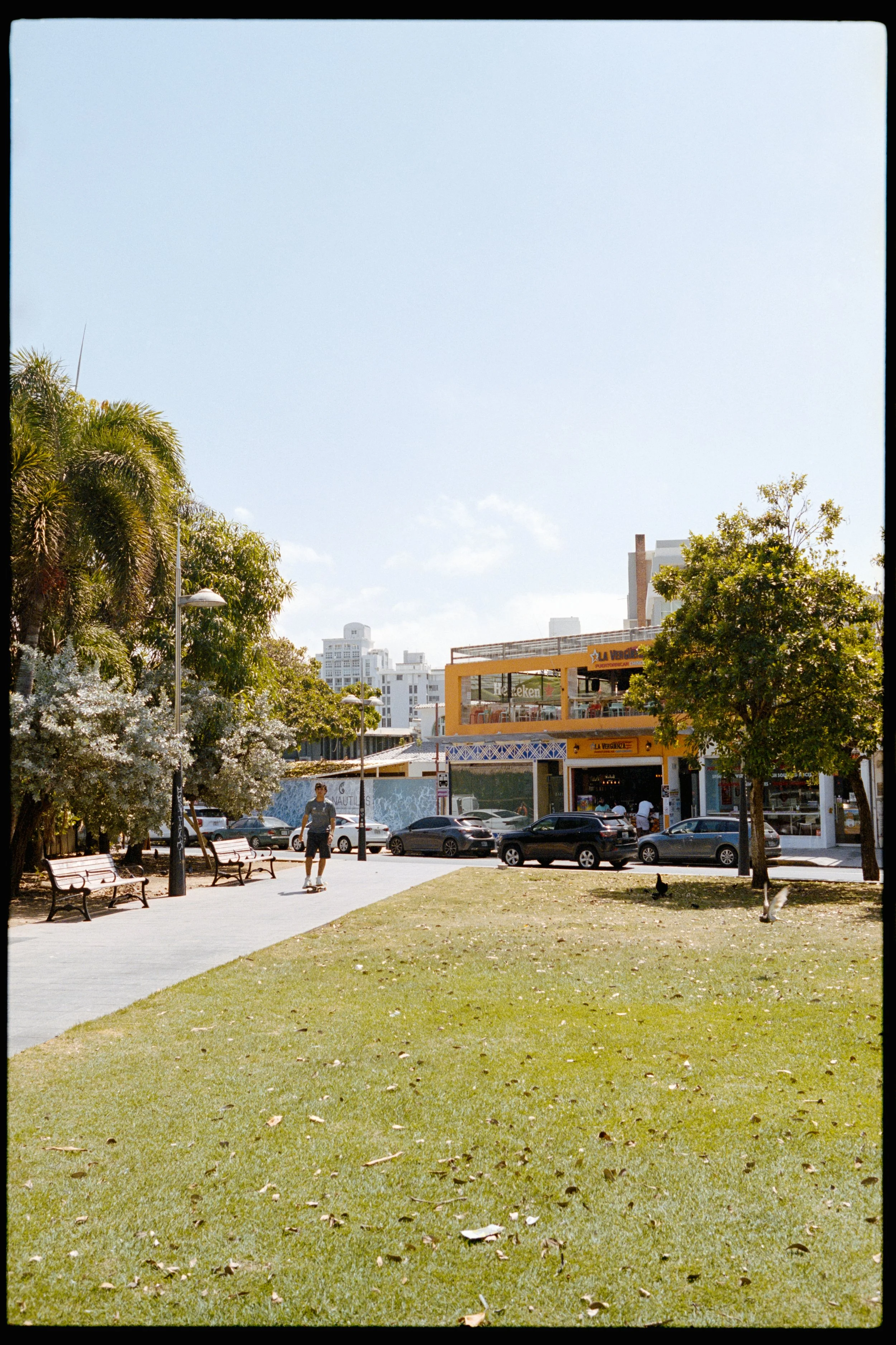 City park with green grass, trees, a paved walkway, benches, and a person skating. In the background, there are buildings and commercial establishments, including a restaurant with a yellow facade and a sign that reads 'La Vereda'.