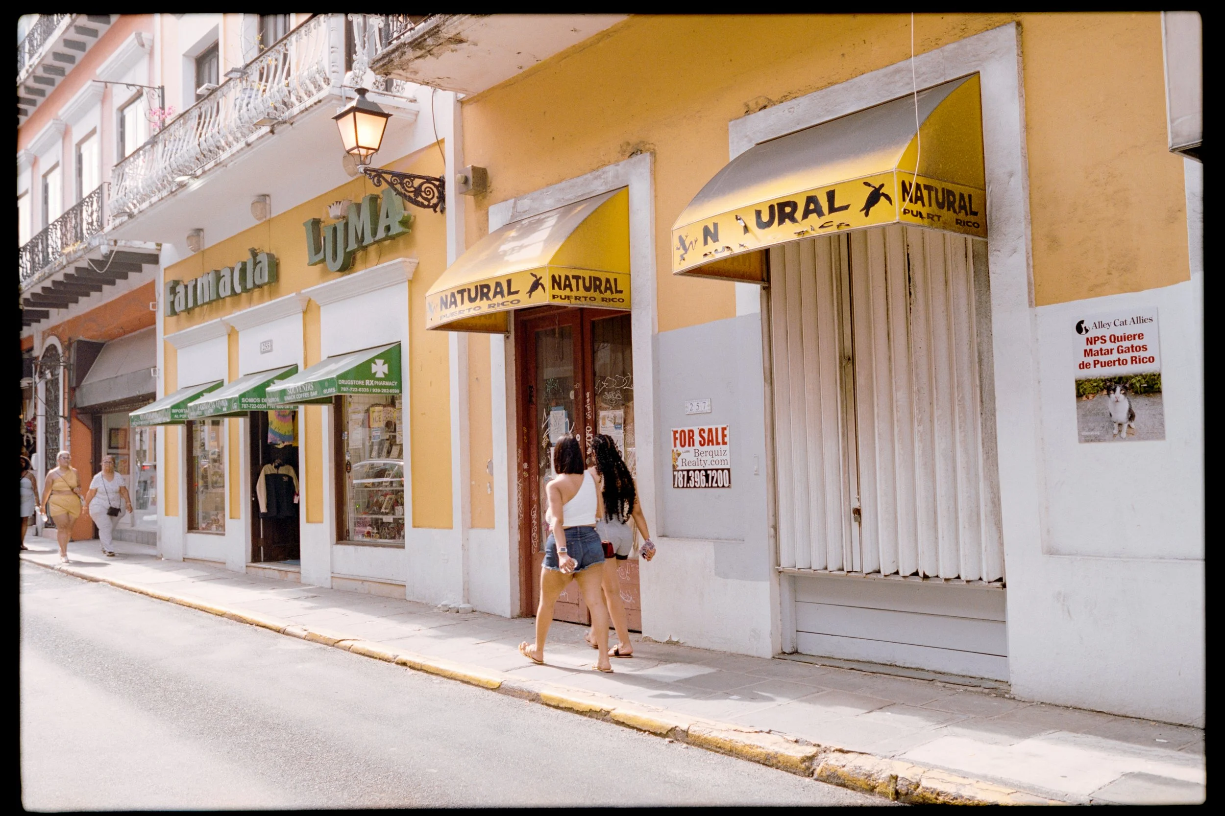 Street view showing a yellow building with storefronts, including a pharmacy and a store with signs reading 'Farmacia Luma' and 'Natural Puerto Rico.' Several pedestrians walk along the sidewalk.