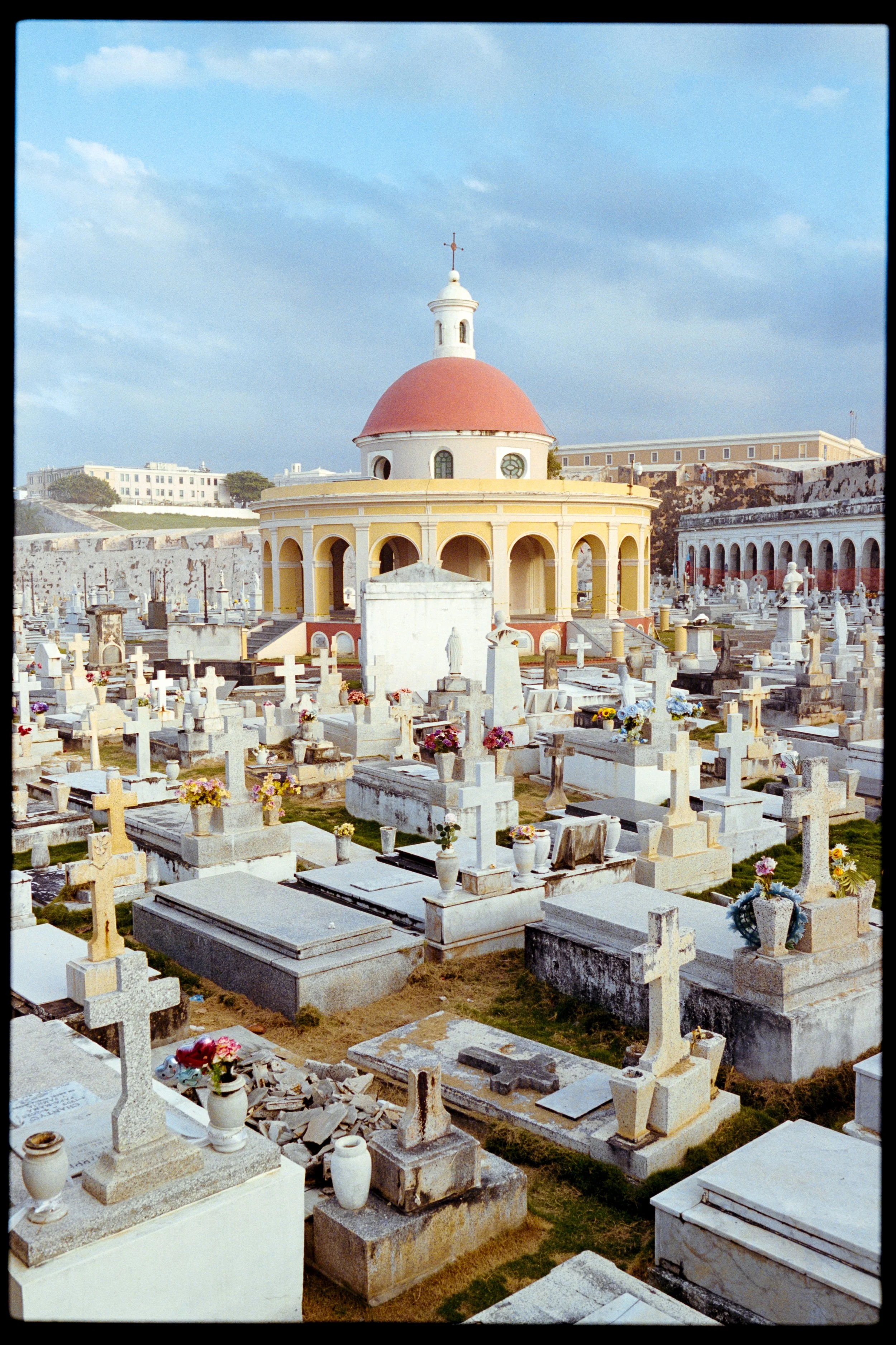 A cemetery with numerous gravestones and monuments, and a small yellow chapel with a red dome and cross on top, set against a cloudy sky.