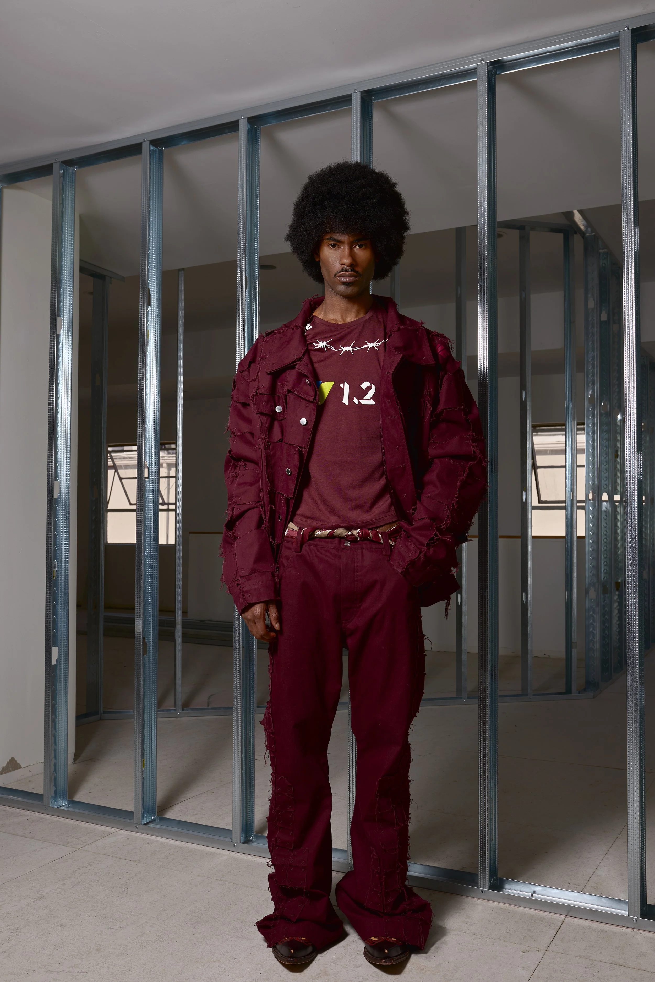 Man standing in front of a metal framing wall under construction, wearing a matching maroon jacket and pants, with a maroon t-shirt and accessories.