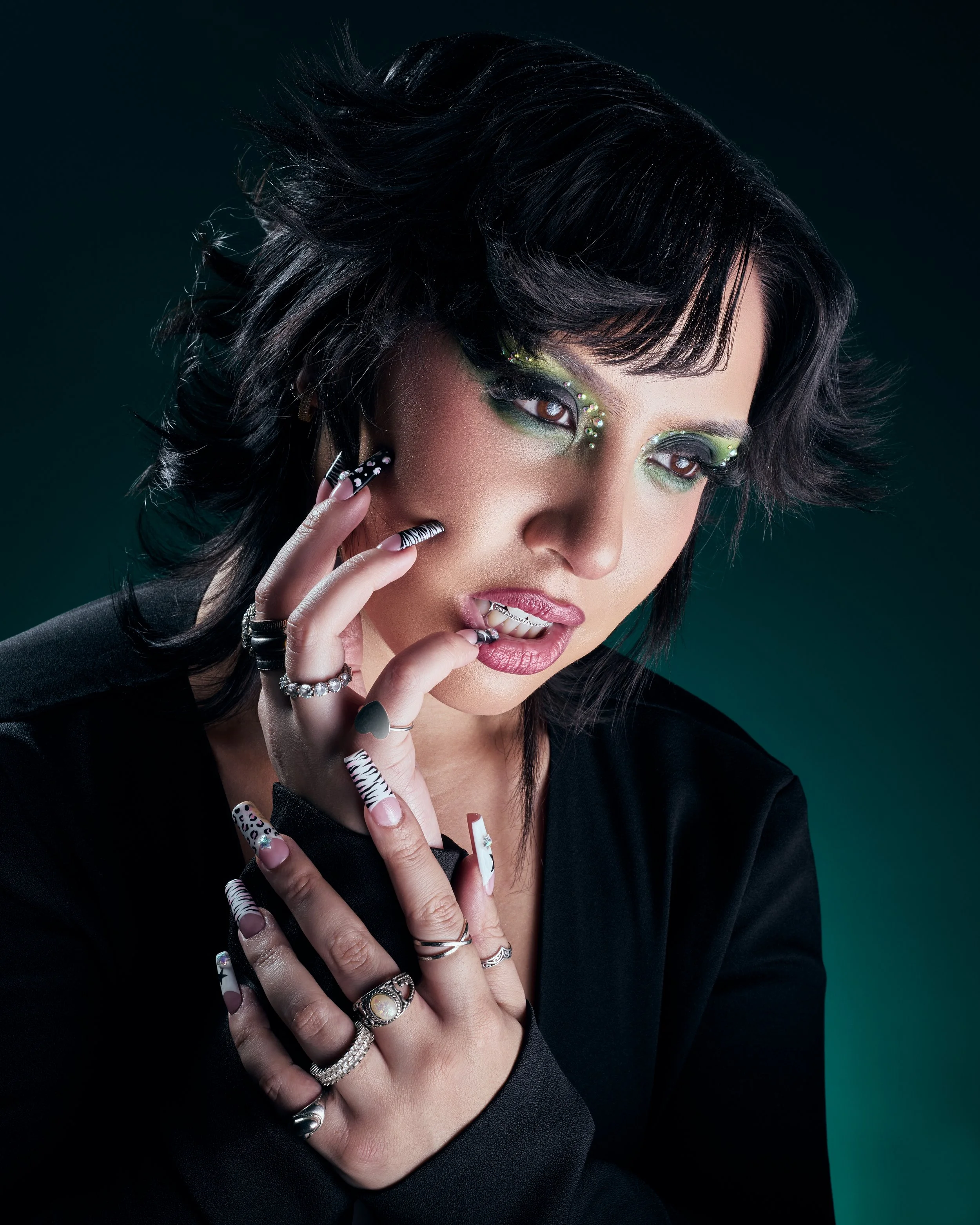 A woman with short black hair and colorful makeup, wearing multiple rings, touching her face and staring thoughtfully.