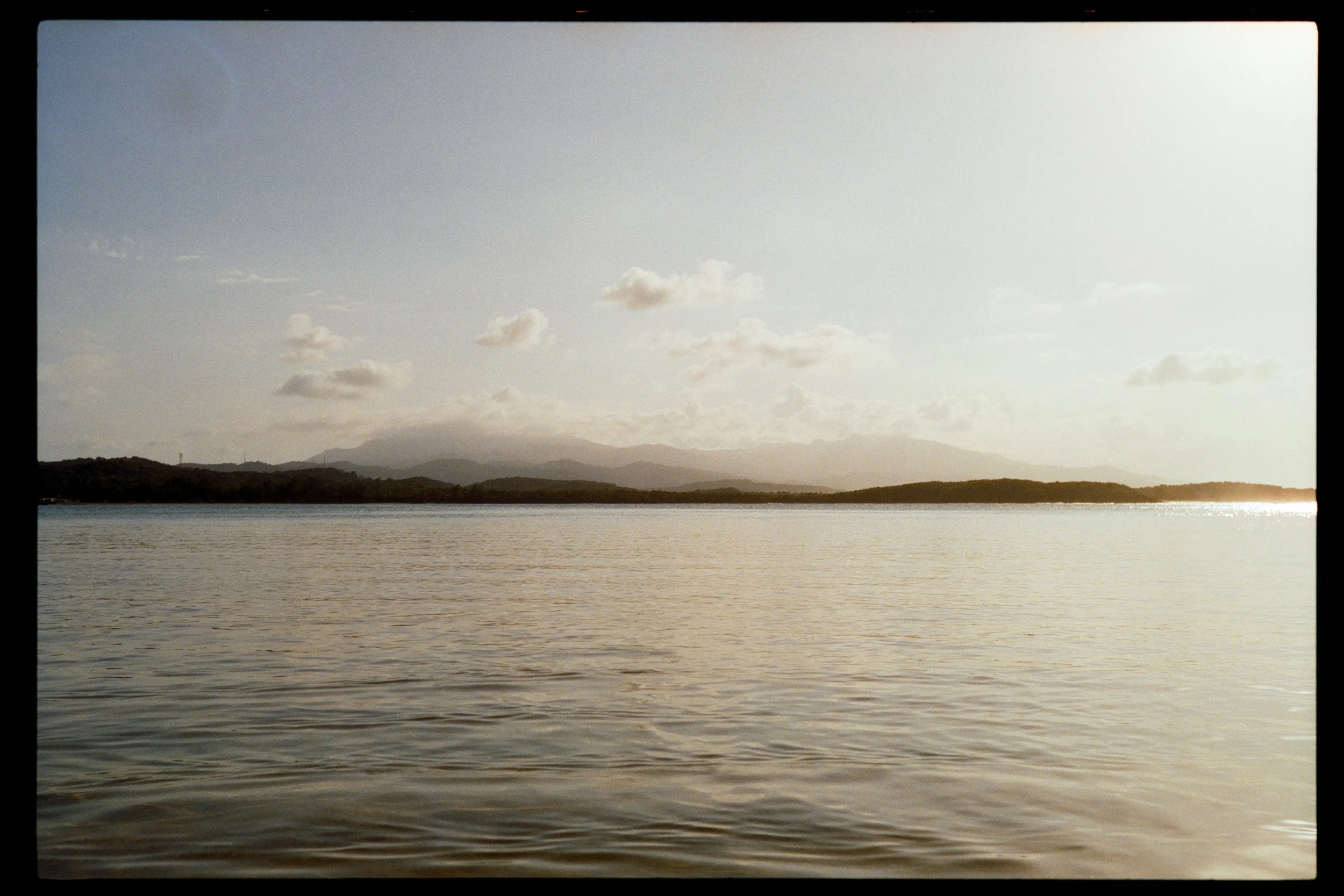 A large body of water with gentle ripples, a distant shoreline with trees and hills under a partly cloudy sky, and sunlight illuminating the scene.