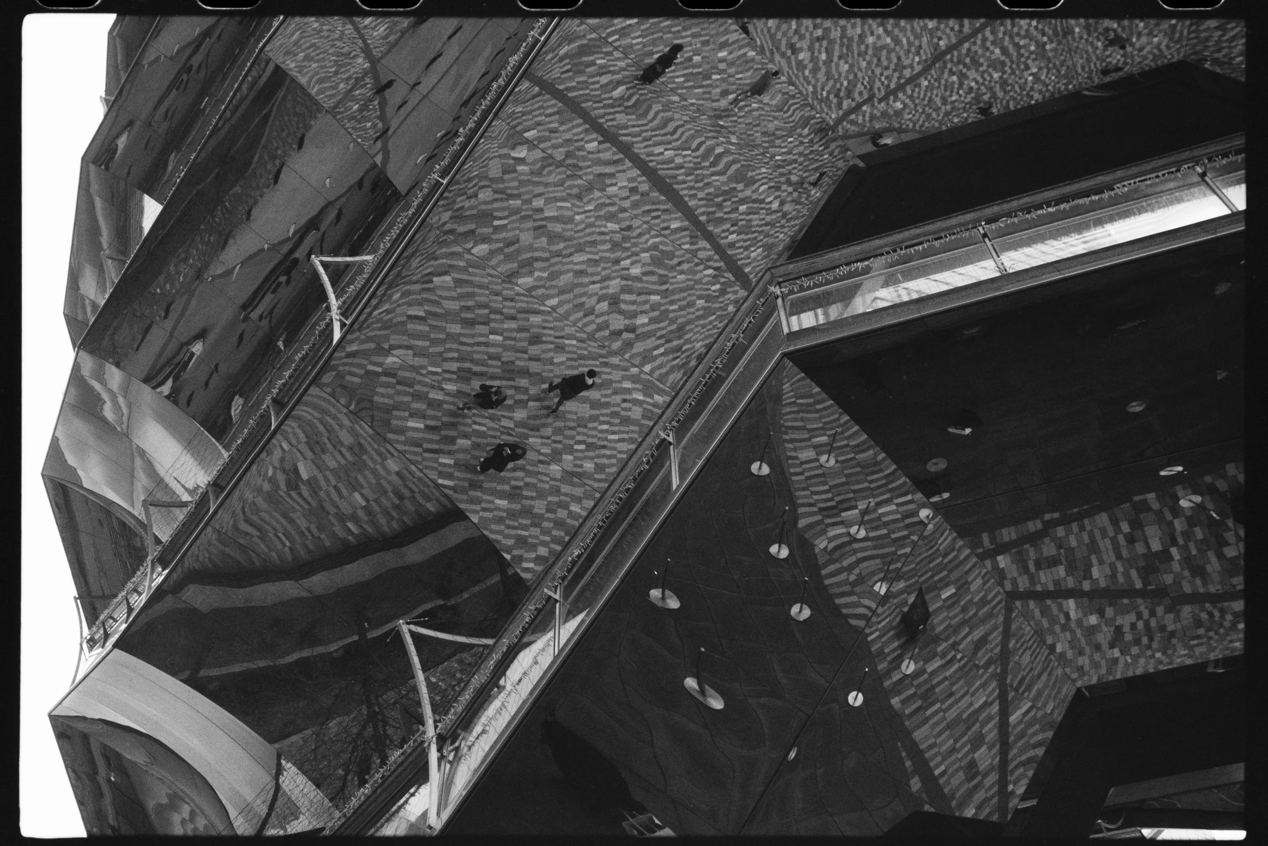 Black and white photo of a building with reflective glass exterior, showing people walking on the patterned pavement below.