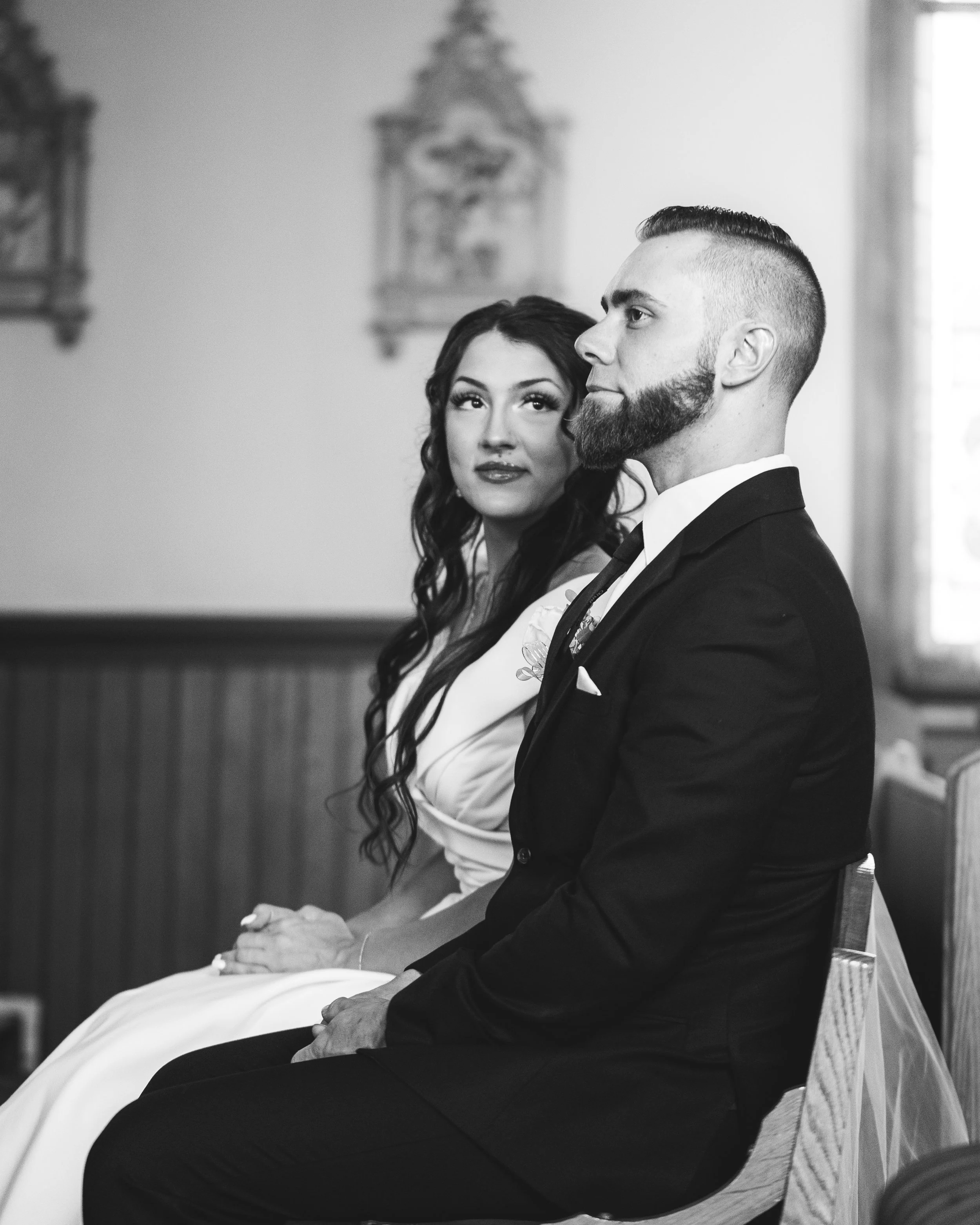 A newlywed couple sitting side by side during their wedding ceremony in a church. The bride has long, wavy hair and is wearing a white dress, while the groom has a beard, short hair, and is dressed in a black suit with a tie.