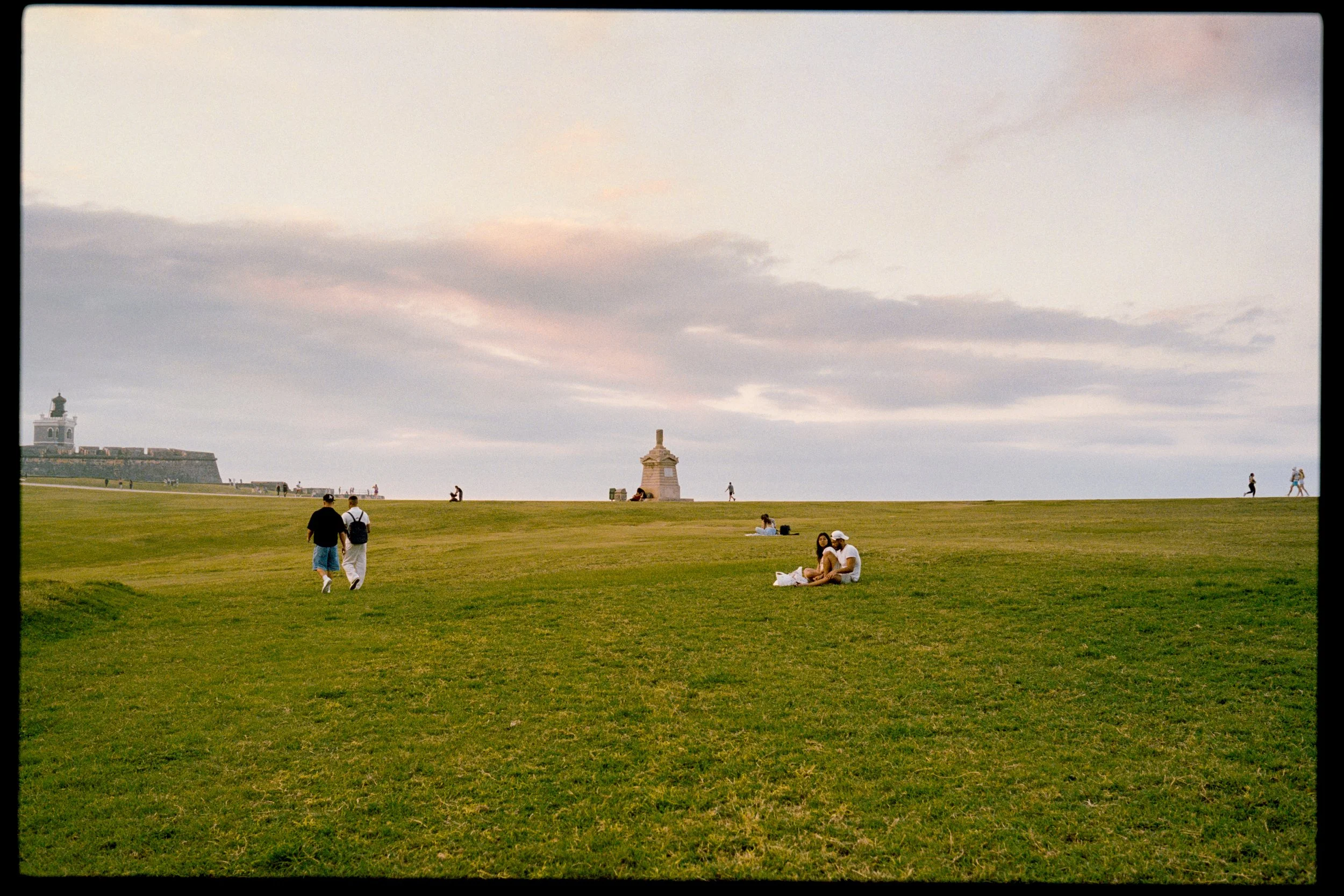 A grassy park with people walking, sitting, and relaxing; a cloudy sky overhead; a lighthouse and castle-like structure in the background.