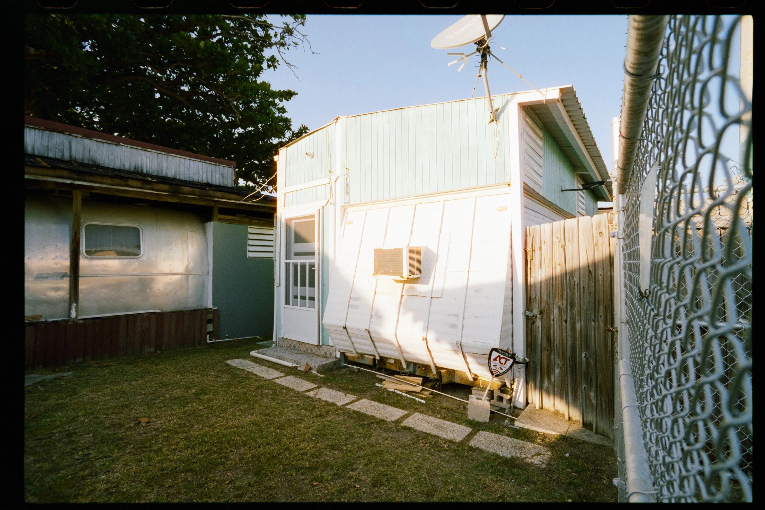 Backyard with a fenced area, small mobile trailer, and a large white storage container or shed with an air conditioning unit attached. A satellite dish is mounted on top of the shed. The yard has grass and stone paving, enclosed by a wooden and chain