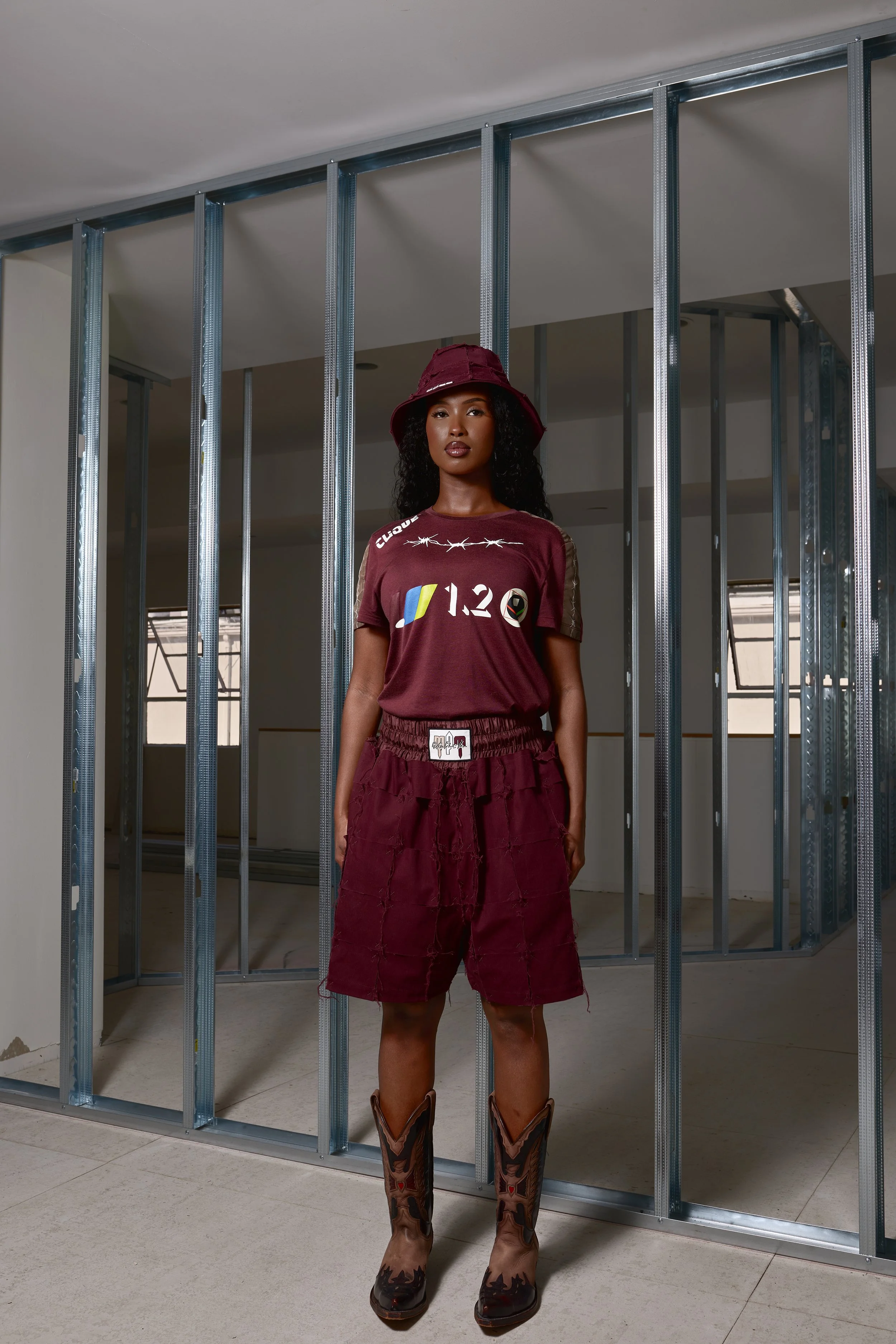 A woman in a maroon outfit, including a bucket hat, t-shirt, shorts, and cowboy boots, standing in an unfinished room with exposed metal framing.