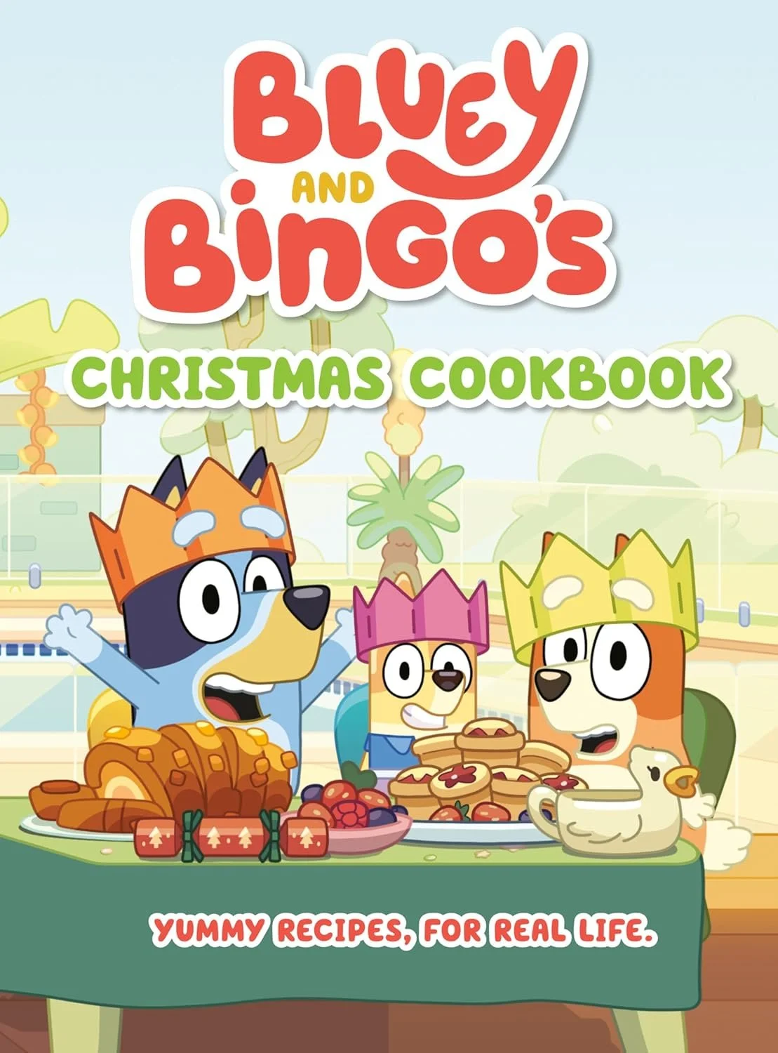 Cover of a children's Christmas cookbook featuring cartoon dogs wearing crowns, with decorated food on a table, set outdoors in a festive scene.