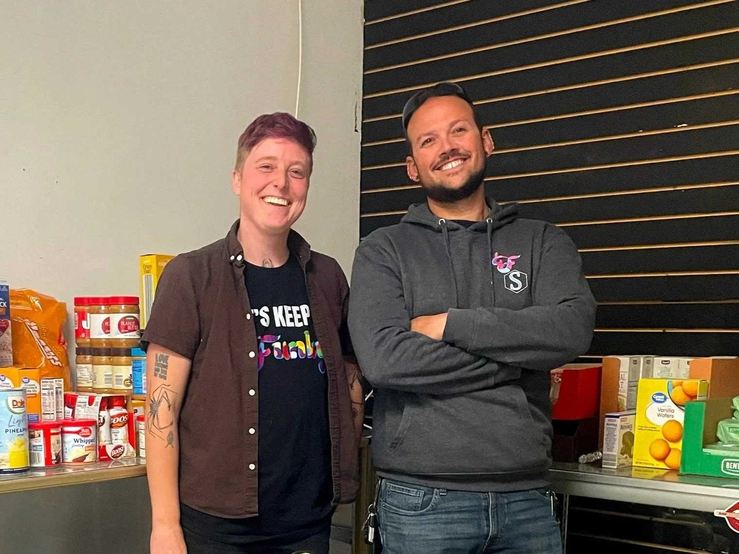 Two people standing in front of a grocery shelf, smiling at the camera. The person on the left is a woman with short hair, tattoos, and wearing a black shirt and bun. The person on the right is a man with beard, wearing a dark hoodie and cap.