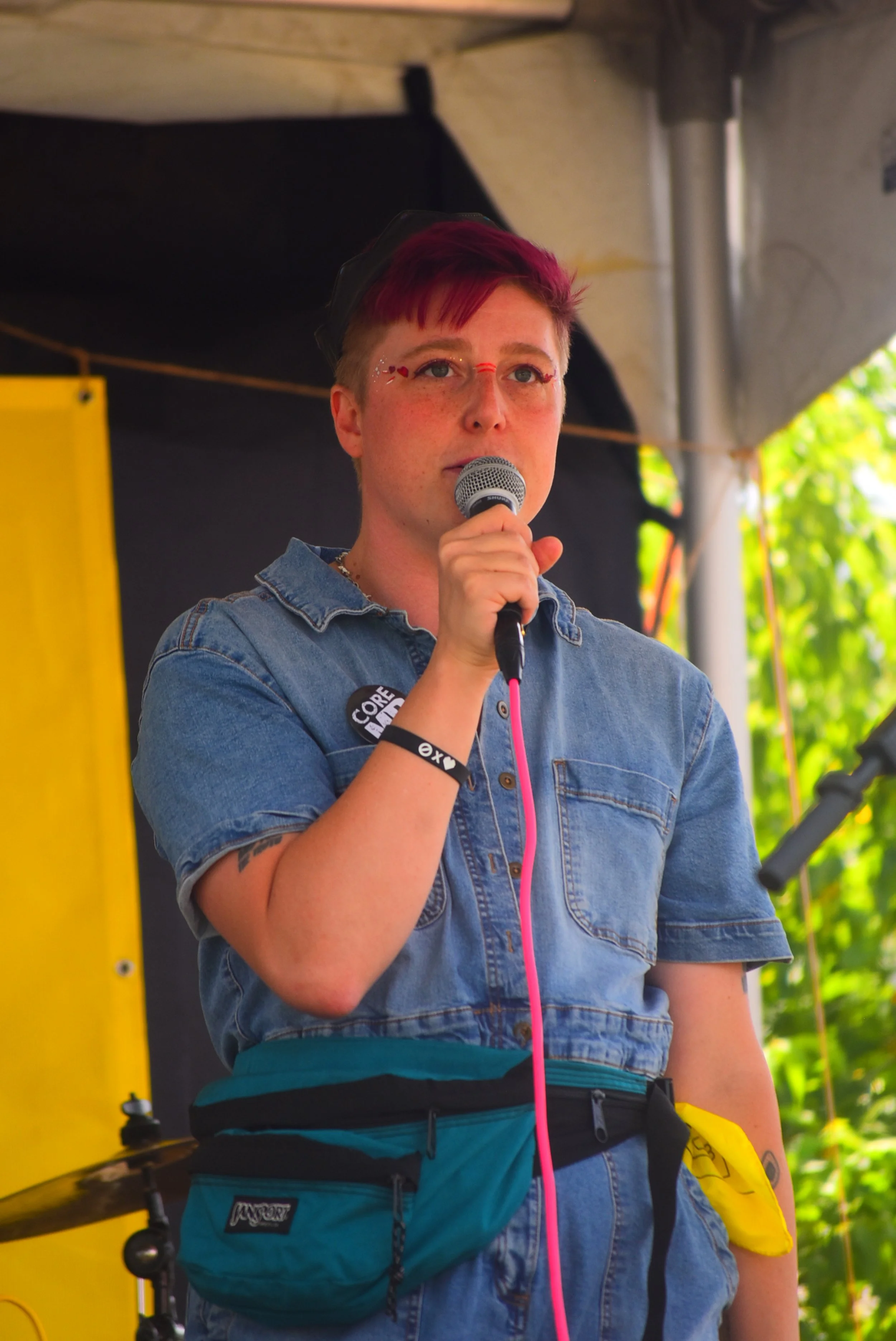 Person with short, red hair speaking into a microphone during an outdoor event, wearing a denim shirt and a teal waist pack.