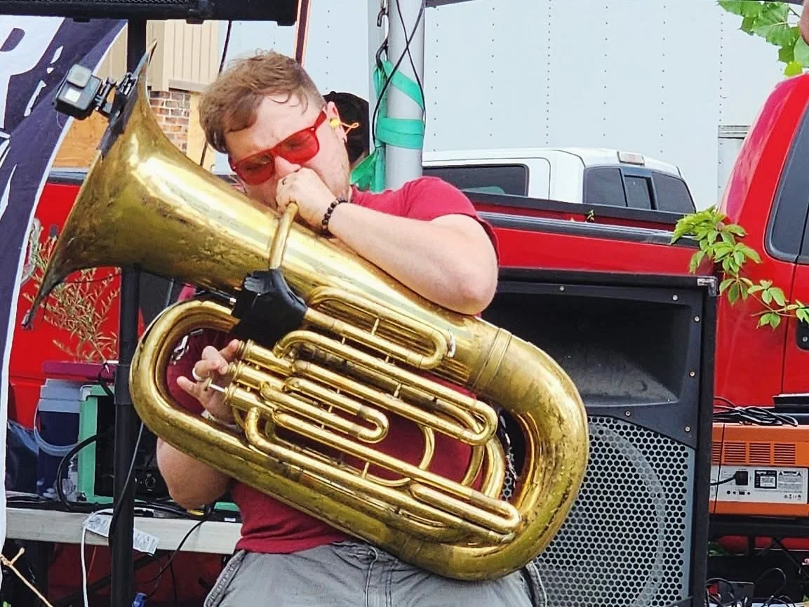 A young man wearing red sunglasses and a red t-shirt playing a large brass tuba at an outdoor event.