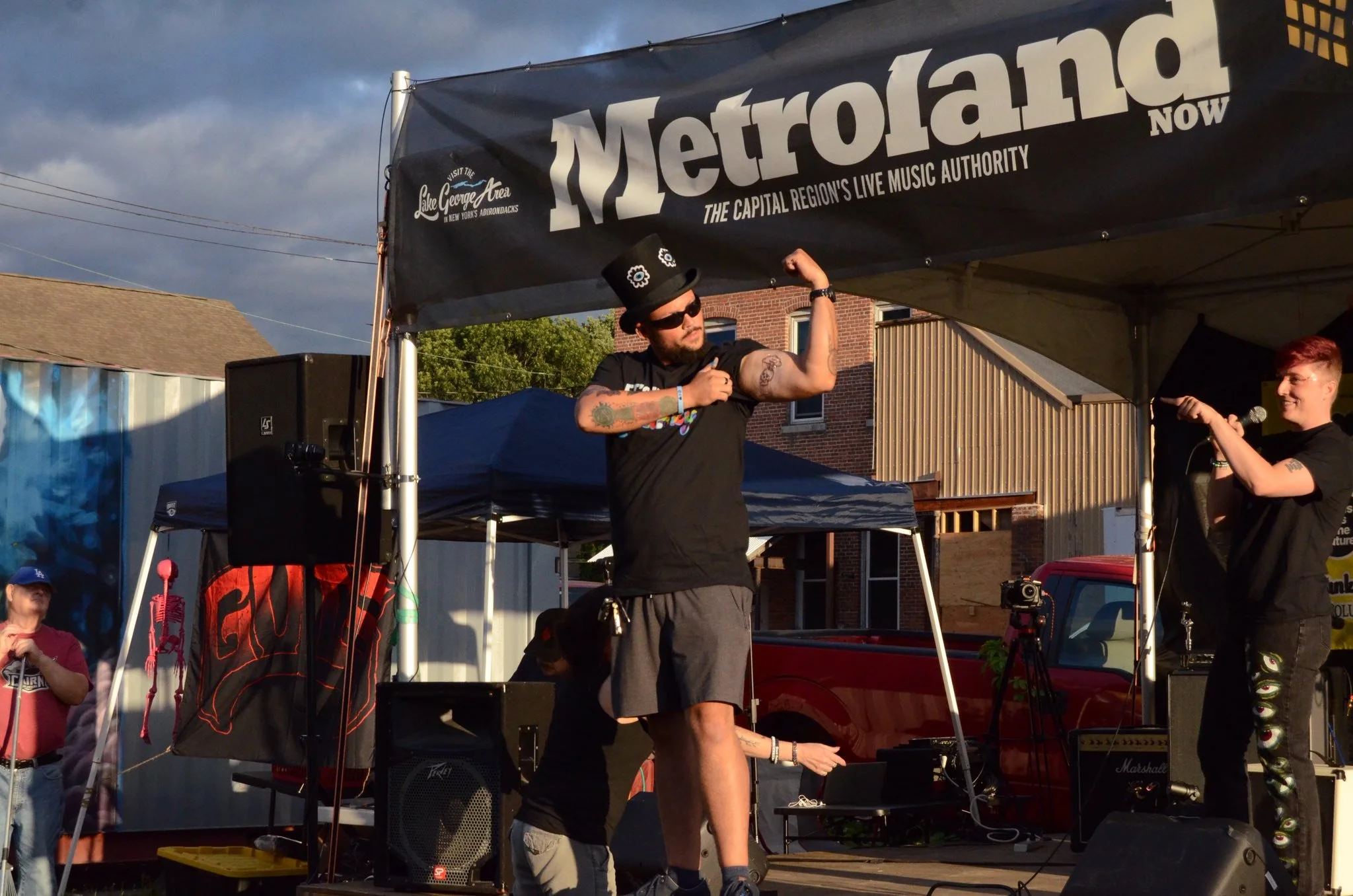 A man with a beard, sunglasses, and a black top hat with gear designs flexes his bicep on a stage at the Metroland music event, with a woman holding a microphone smiling nearby. The stage has a black banner with the text 'Metroland' and 'Lake George Area' and is set outdoors with sunlight and dark clouds overhead.