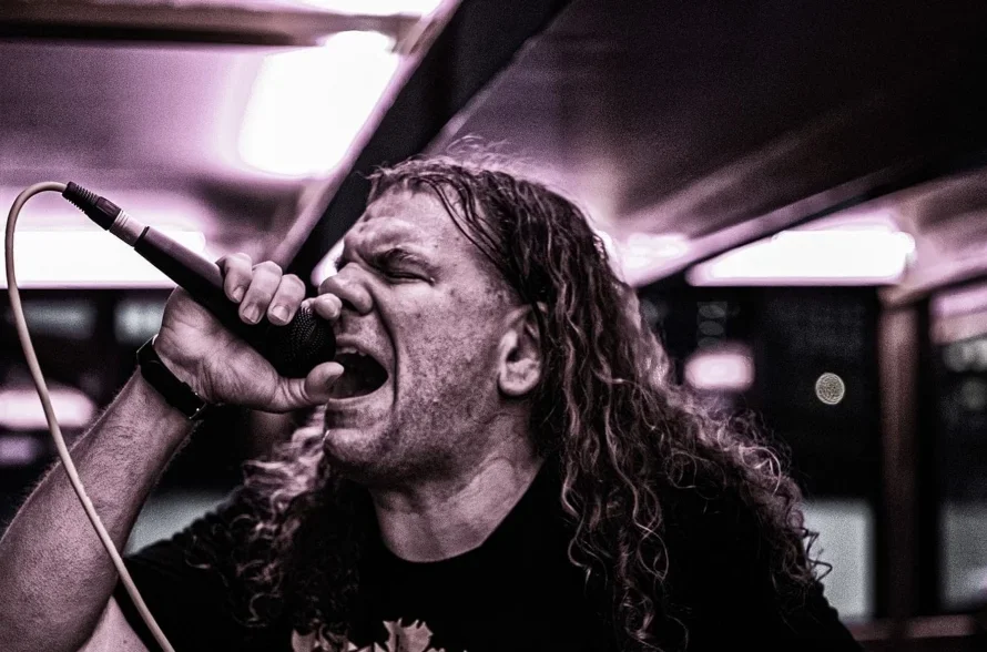 A man with long, curly hair singing passionately into a microphone in a dimly lit setting.