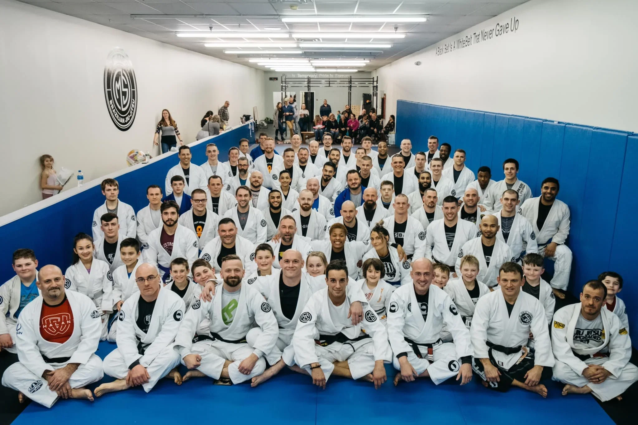 Group photo of martial artists in white gis with black belts, sitting and standing on blue mats in a gym, with spectators in the background.