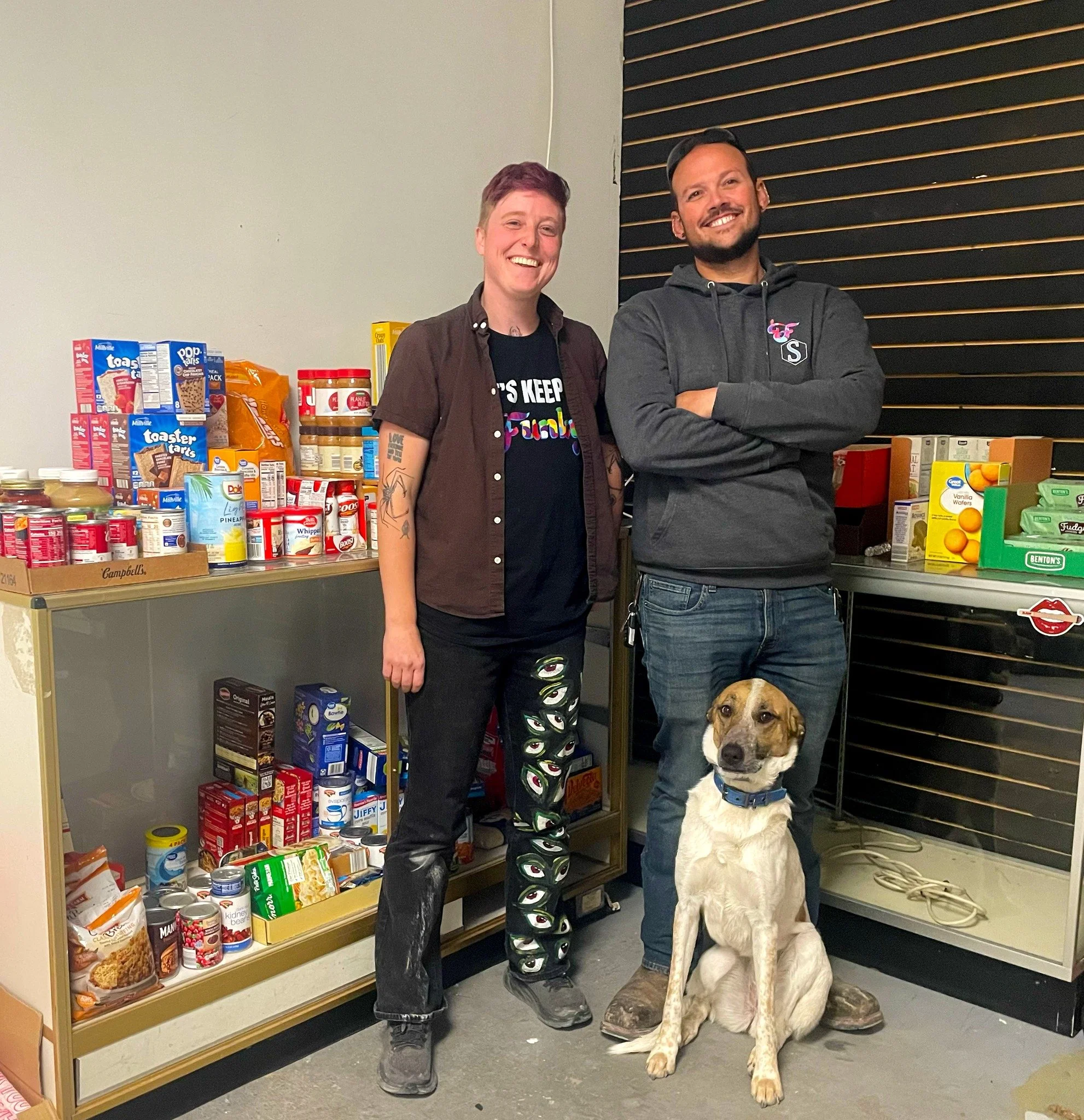 Two people and a dog standing in a room with shelves of canned and packaged food. One person is wearing a black shirt and brown shirt, the other in a dark hoodie with arms crossed, and the dog is sitting in front of them.
