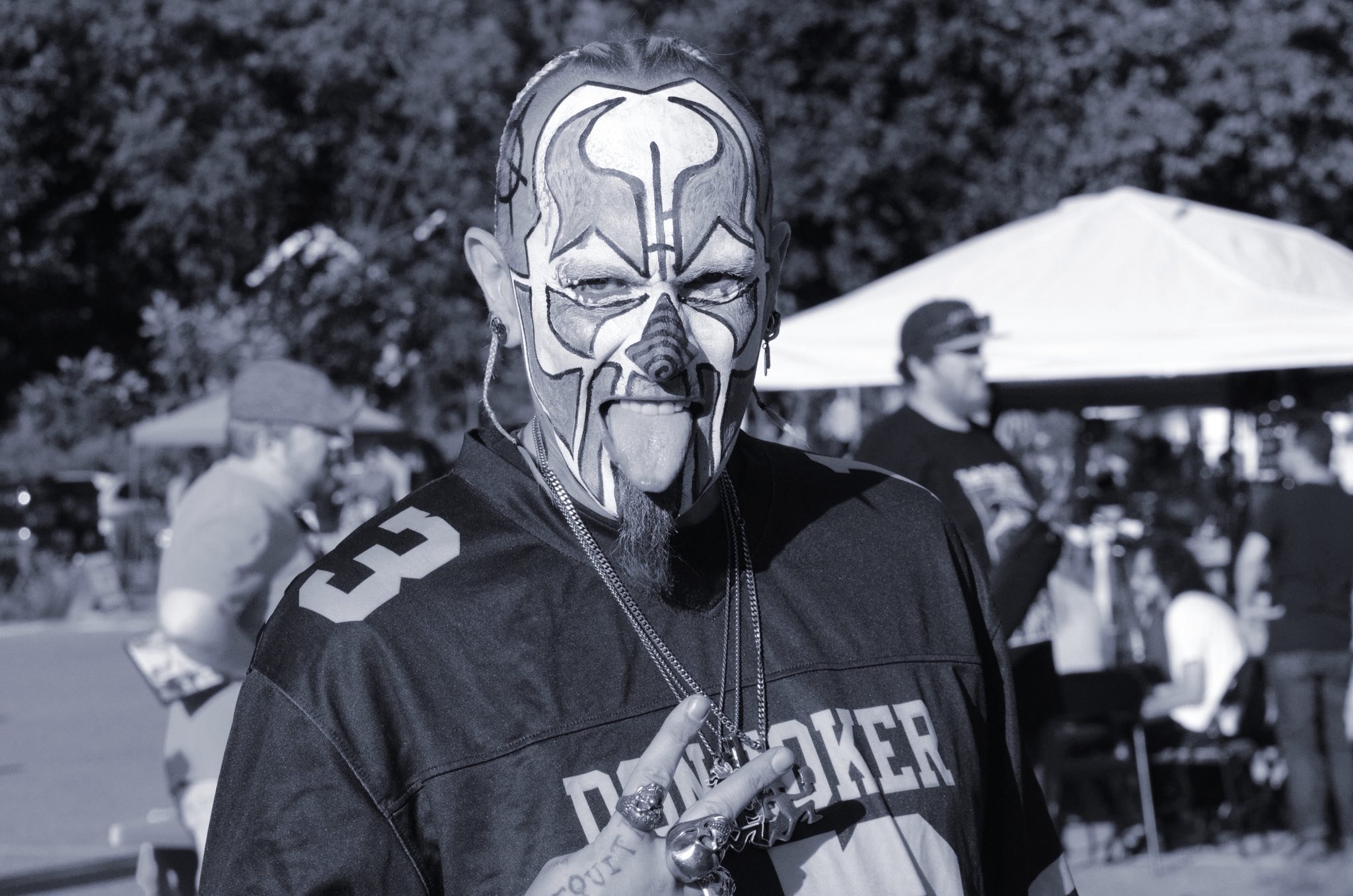 A man with face paint, wearing a sports jersey, making a peace sign at an outdoor event
