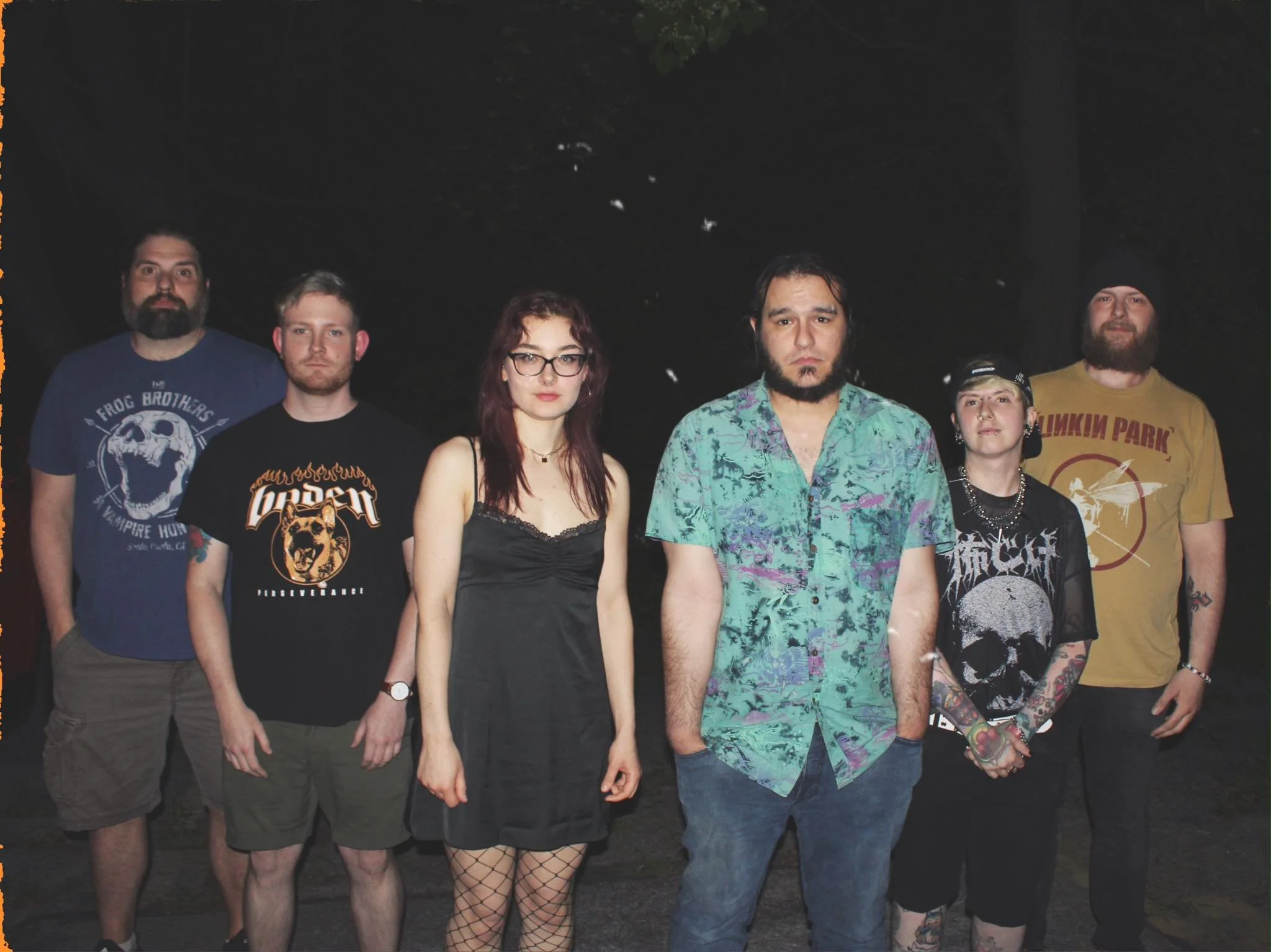 Group of seven people standing outdoors at night, dressed casually, with some wearing band t-shirts, in front of dark trees.
