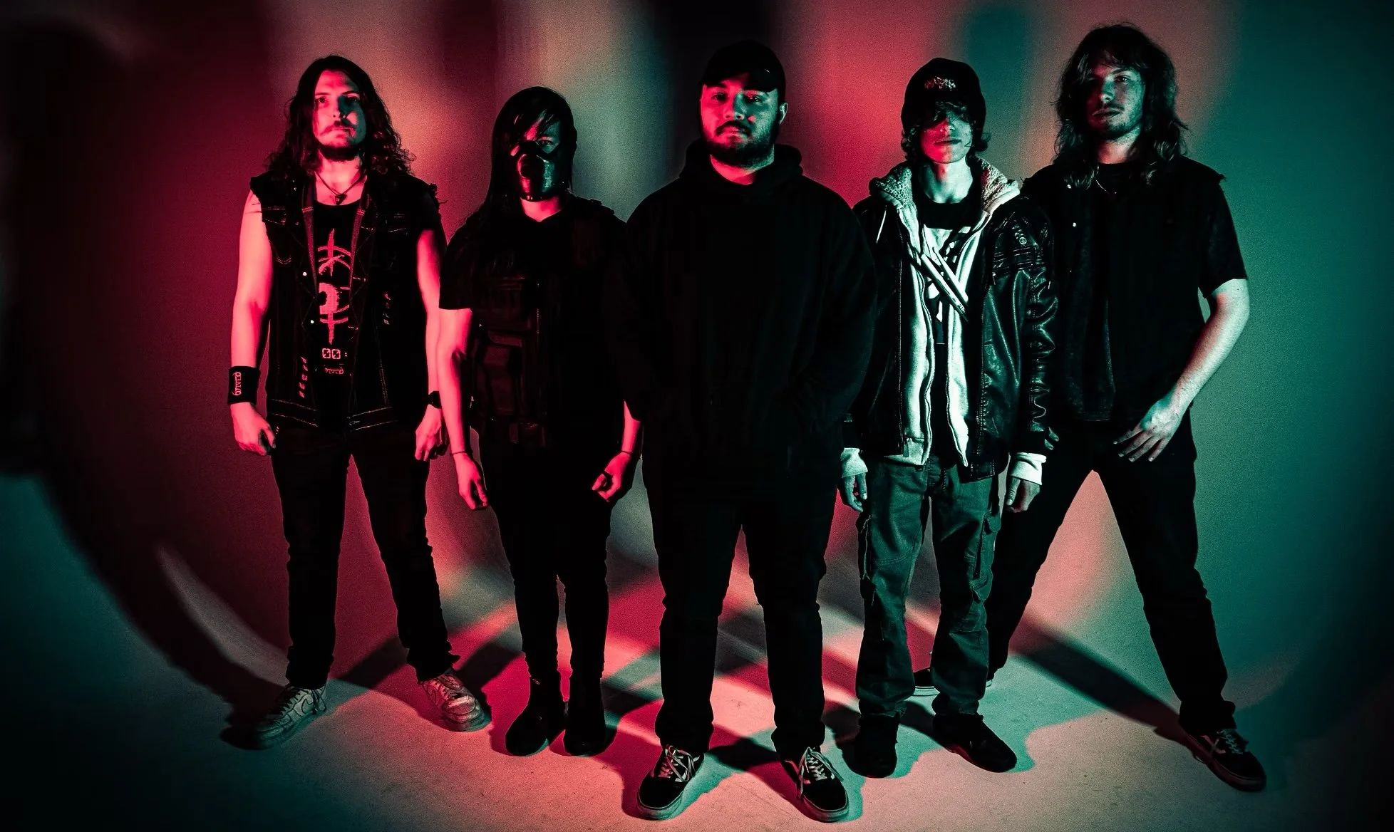 A group of five young adults standing in a dimly lit room with colorful lighting, dressed in edgy, casual, rock-inspired clothing.