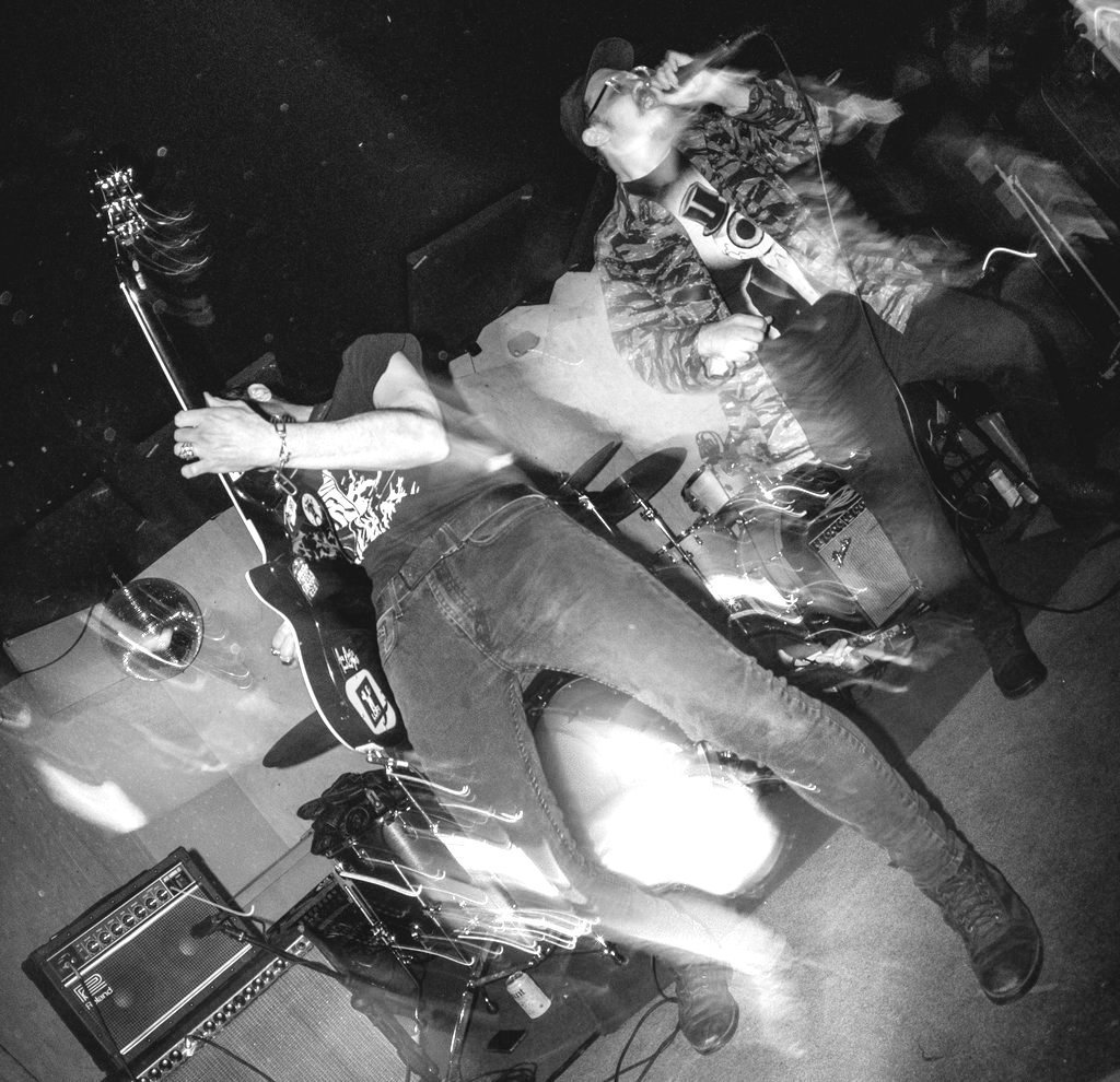 Black and white photo of a live music performance with two musicians on stage, one playing guitar and the other singing into a microphone, with musical equipment around them.