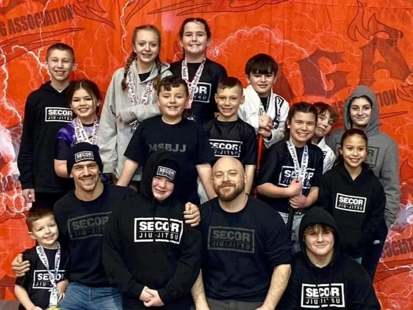 Group photo of children and adults at an indoor event, some wearing black T-shirts with 'SECOR JIU-JITSU' logo, against a red backdrop with lightning graphics.
