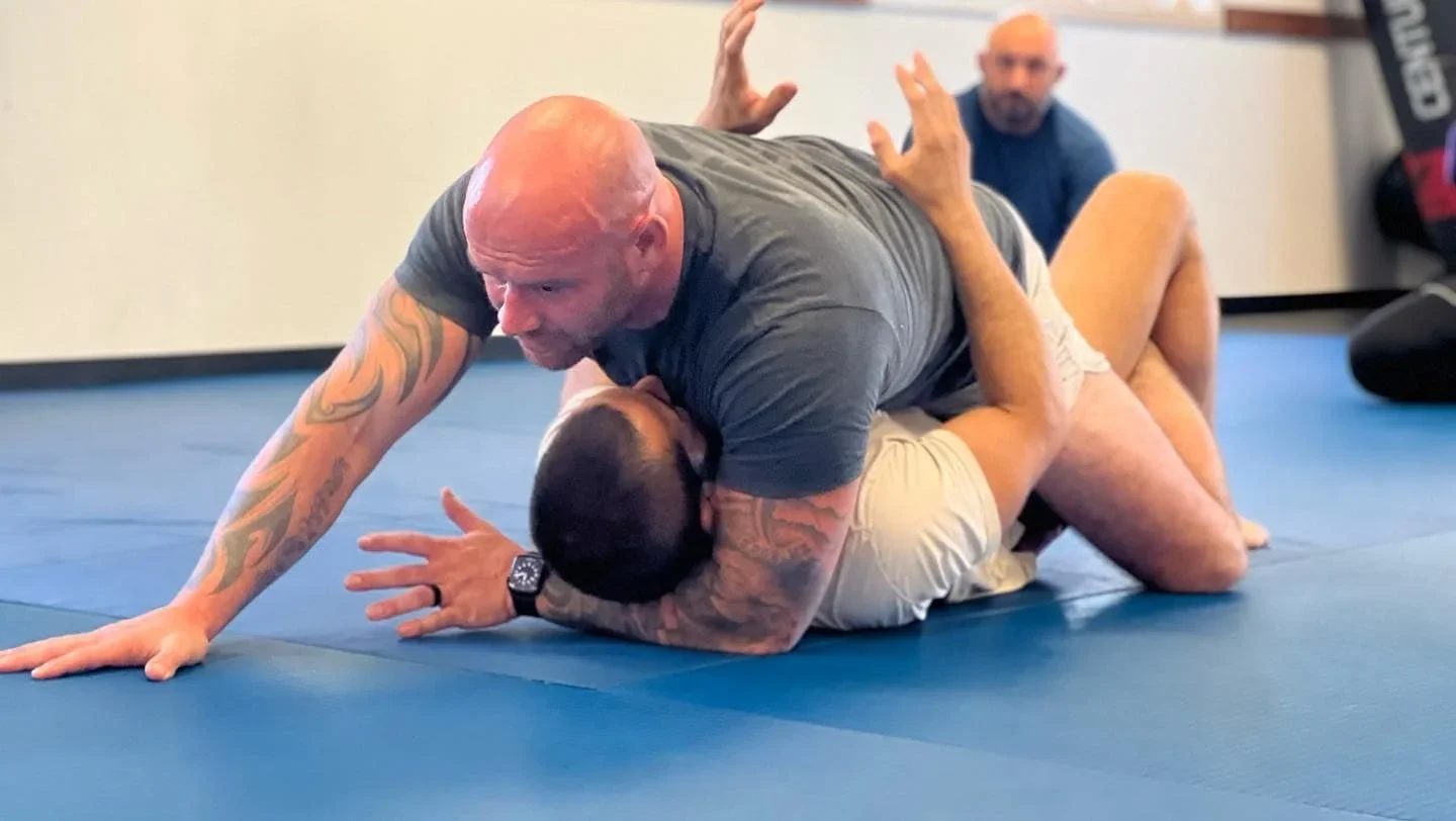 Two men practicing Brazilian Jiu-Jitsu on a blue mat, with one man on top attempting a choke hold.