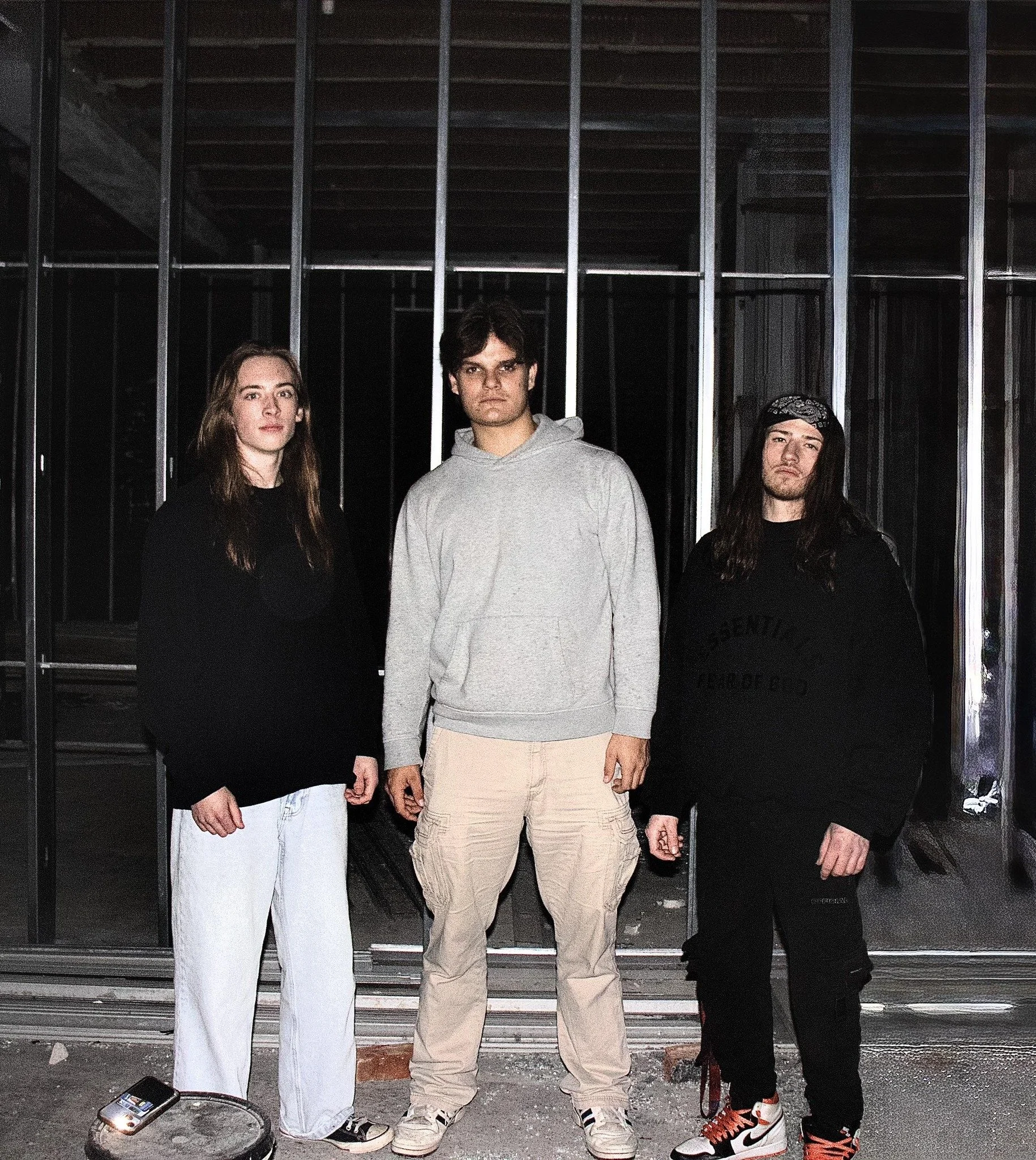 Three young adults standing in front of a construction site with metal framing. They are wearing casual clothing, with the two women on the left and right in black and white, and the man in the middle in a light gray hoodie and beige cargo pants.
