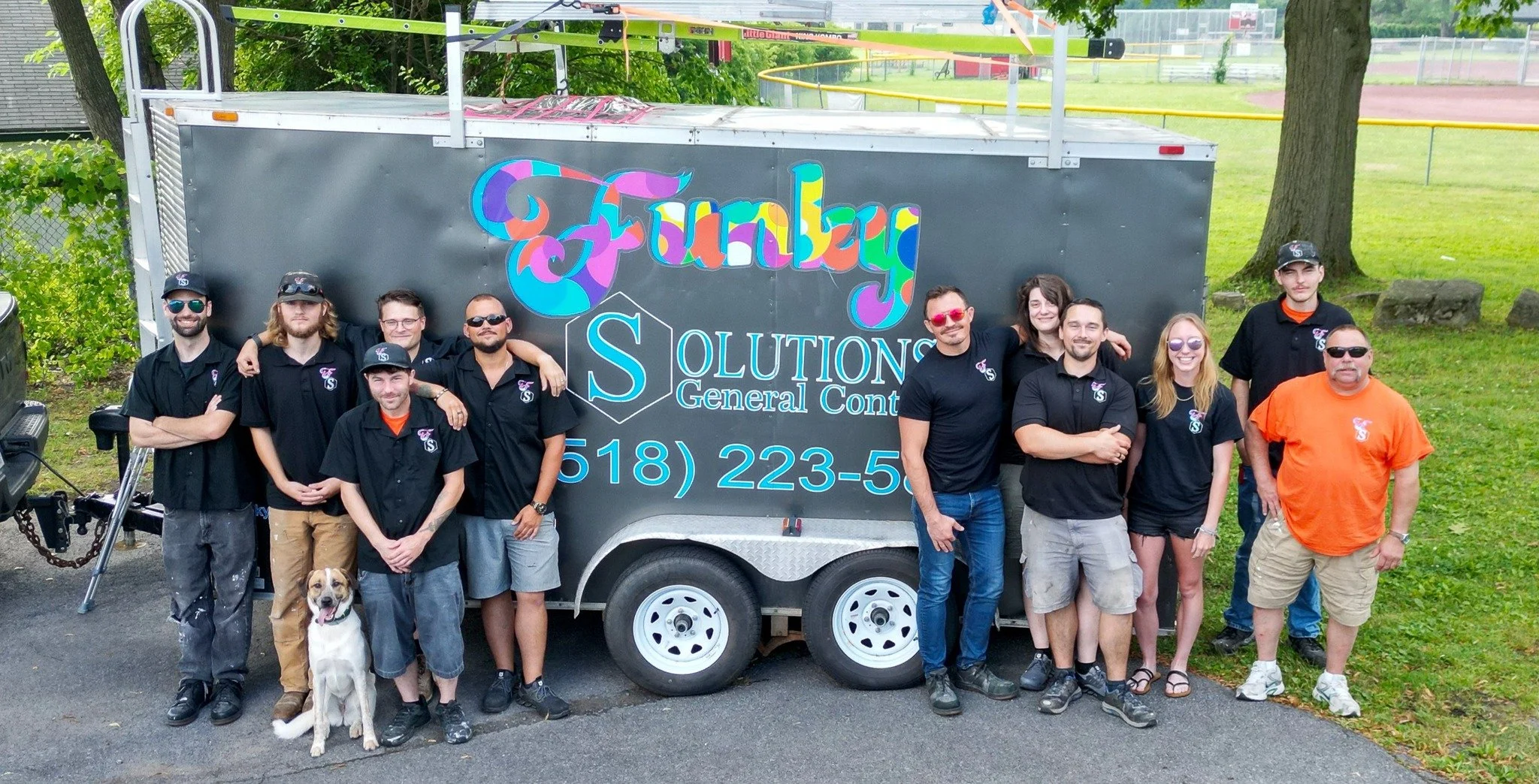 Group of people standing in front of a trailer with a sign that reads "FAMILY SOLUTIONS GENERAL CONTRACTING" and a phone number, outdoors on a paved area with grass, trees, and a sports field in the background.