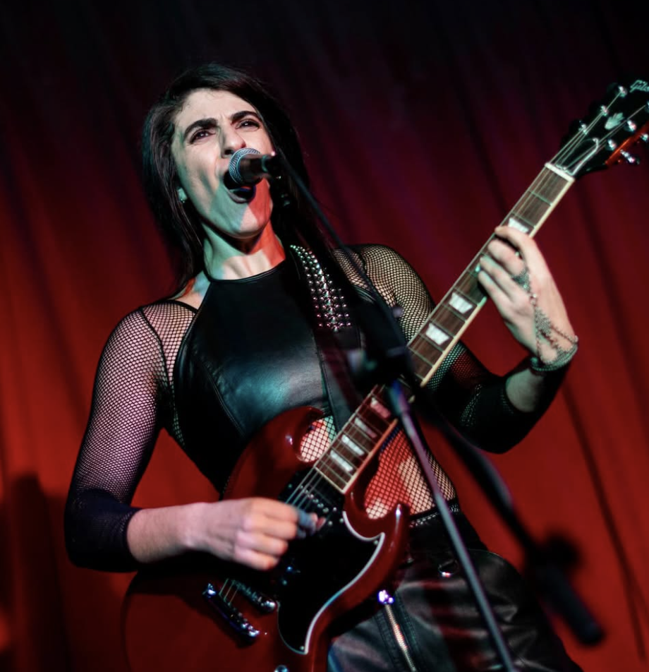 Female musician performing on stage with a guitar and singing into a microphone, red stage curtain in the background.