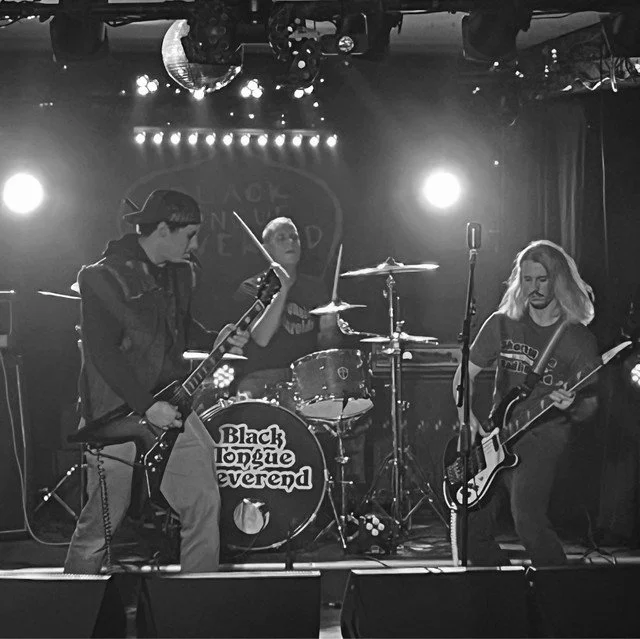 A black and white photo of a three-member rock band performing on stage with drums, electric guitars, and microphones.