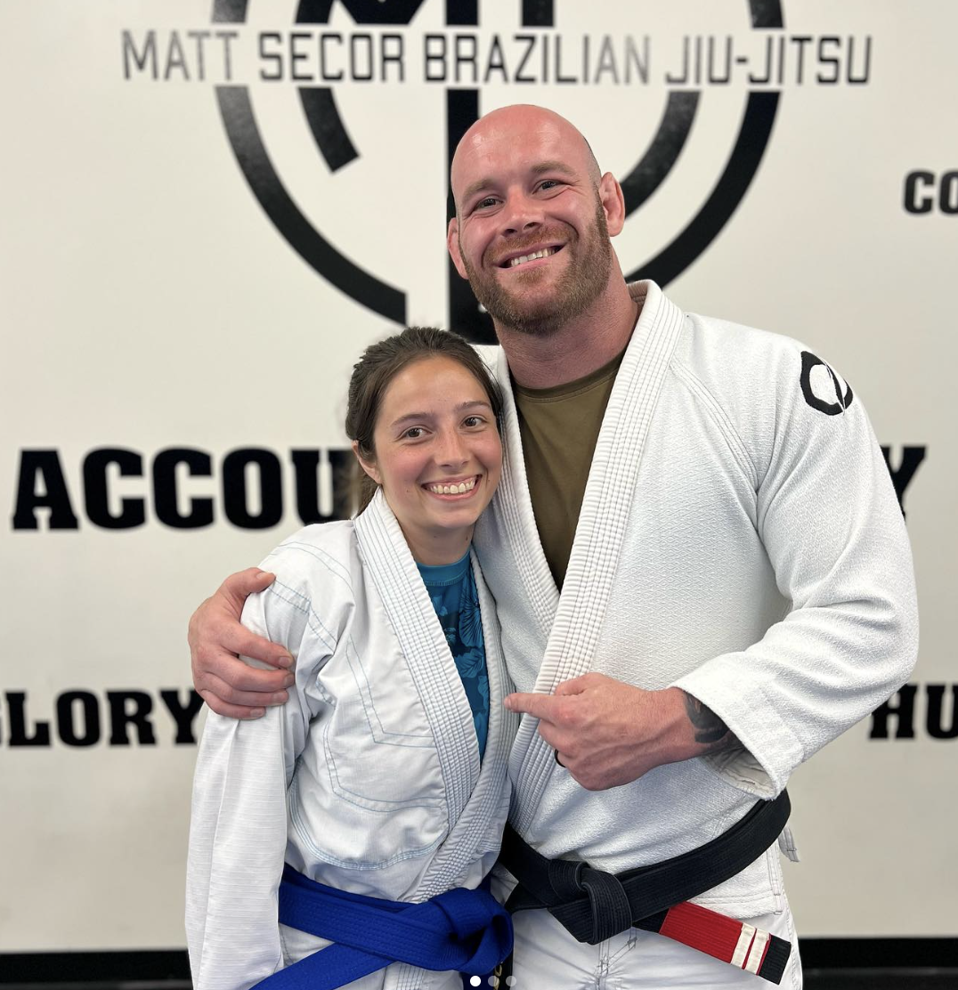 Two people in martial arts gis, a woman with a blue belt and a man with a black belt, smiling and posing together in a martial arts academy.