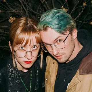 A young woman with glasses and red hair, and a young man with glasses and teal-colored hair, posing closely together.